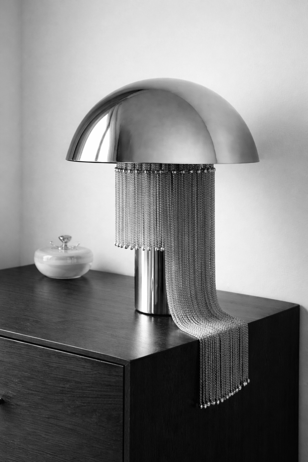 A modern, metallic table lamp with a dome-shaped shade and hanging chain-like decoration on a wooden surface.