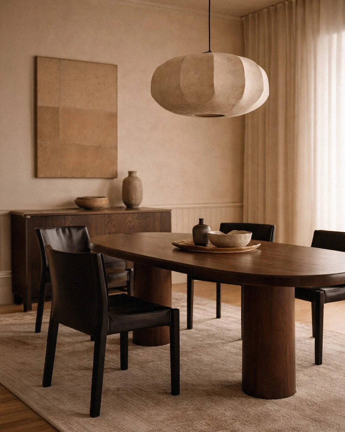 A minimalist dining room with a wooden oval table, black chairs, and a beige rug. There is a large hanging light fixture above the table and simple vases and bowls on the table and a sideboard. Curtains are partially visible near a window.