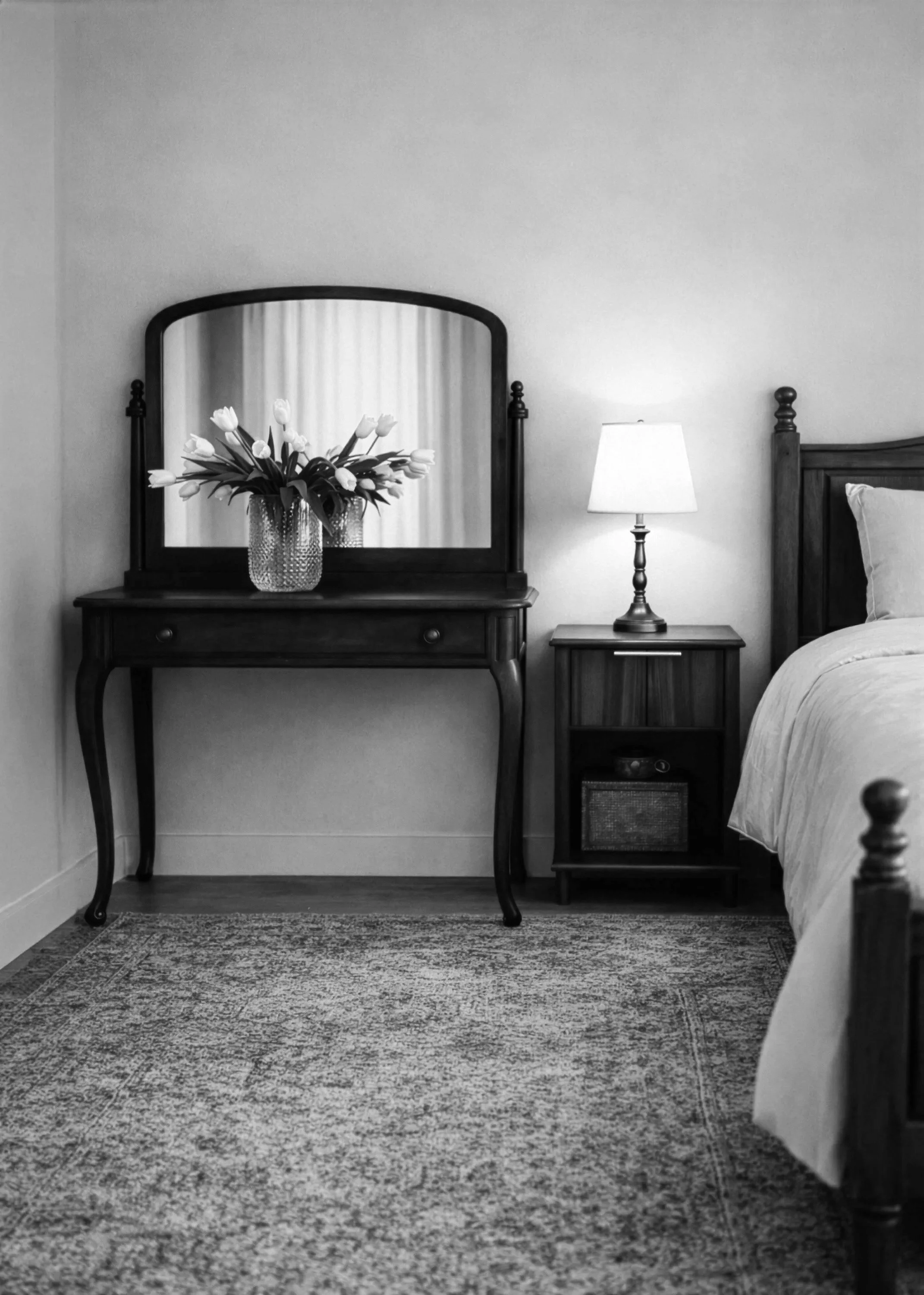 A bedroom with a wooden dresser and mirror, a vase of white tulips, a bedside table with a lamp, and part of a bed with a pillow, all in black and white.