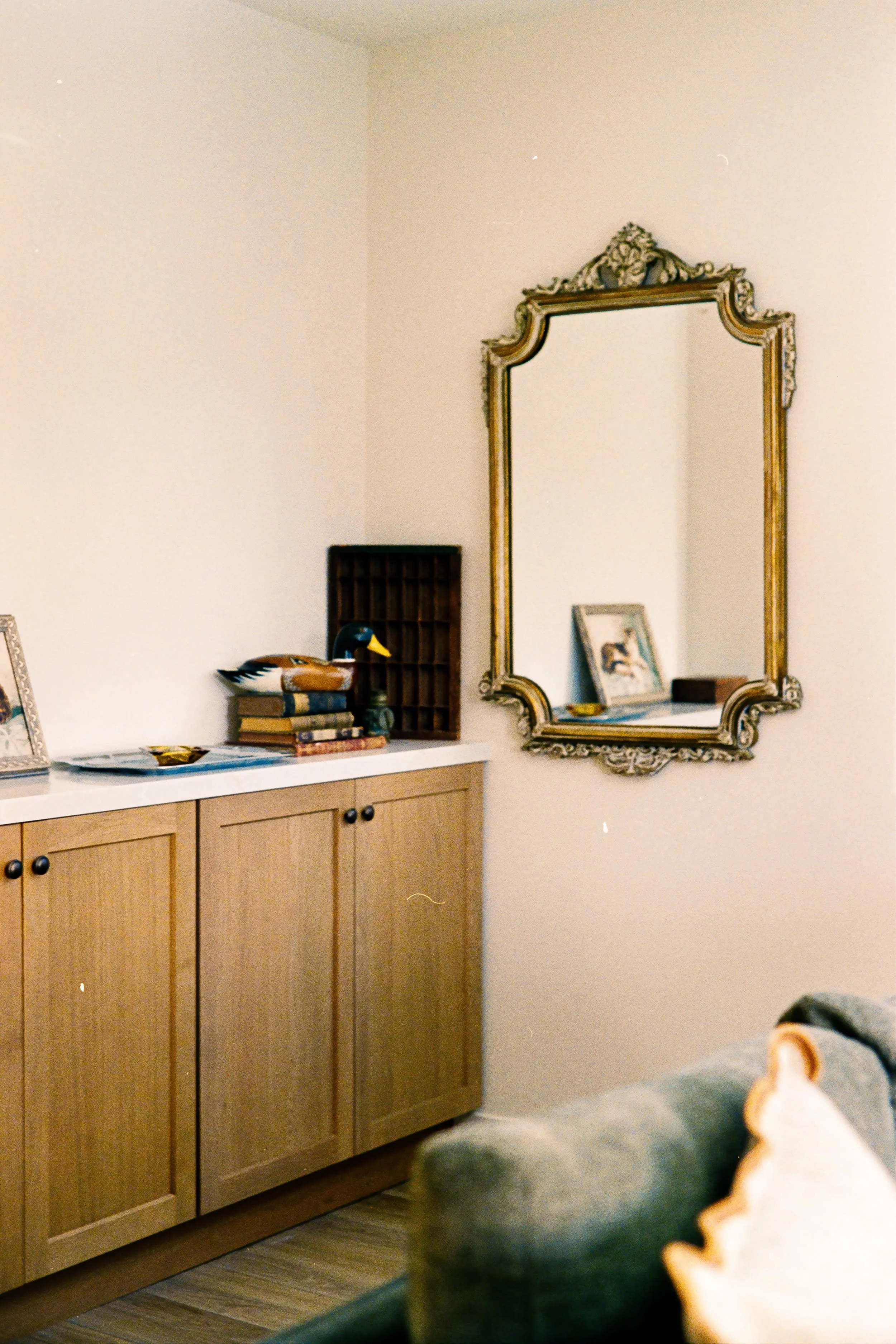 A bedroom corner with a large ornate mirror on the wall, a wooden cabinet with books and decorative items, and a framed picture on top.