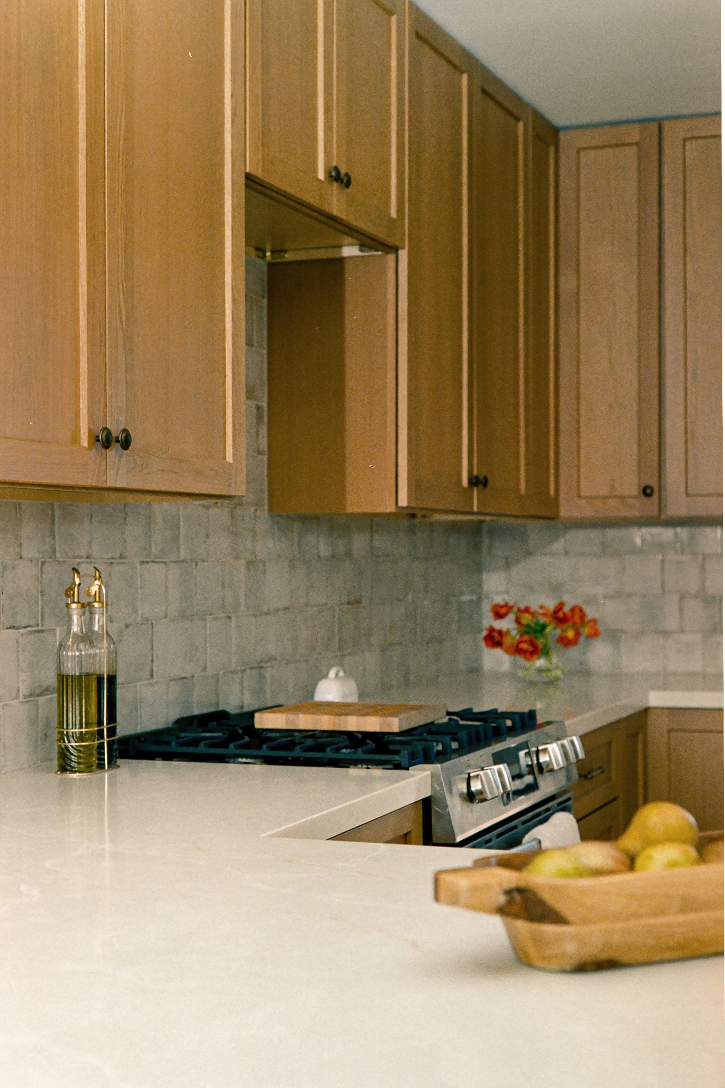 A kitchen with wooden cabinets, a beige countertop, a gas stove, a bottle of oil, a wooden bowl with apples, and a vase with red flowers.