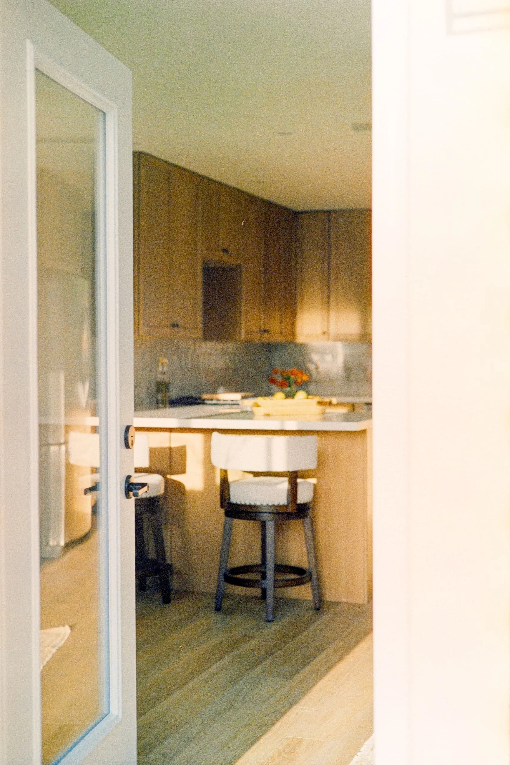 View through a glass-paneled door into a kitchen area with wooden cabinets, a small counter with a bowl of fruit, and two chairs with dark wood frames and cushioned white seats in a cozy dining nook.