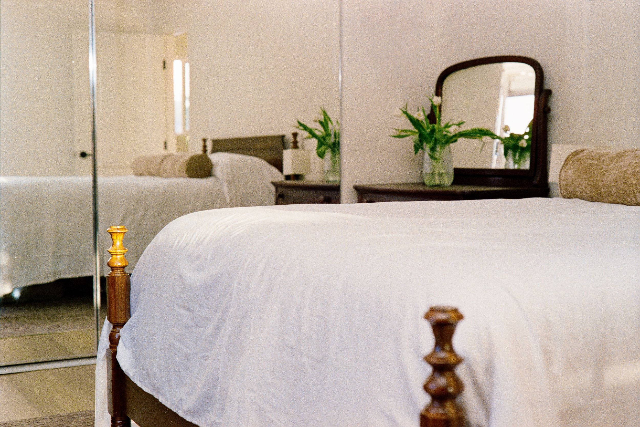 A bedroom with two twin beds, white bedding, a wooden headboard, and a bedside table with a vase of white flowers and a mirror. There is a mirror on the closet door reflecting the room.