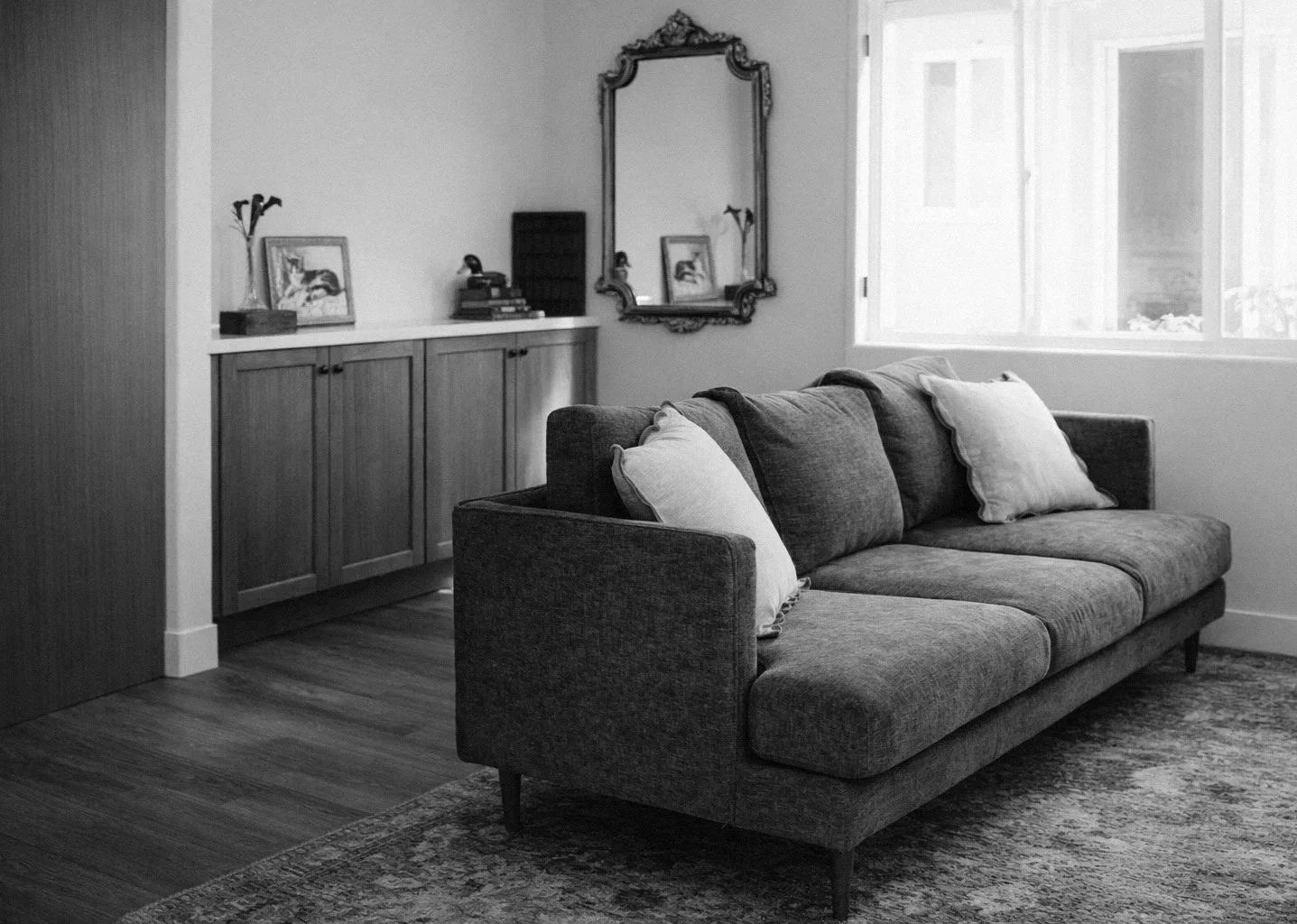 A black and white photo of a living room with a couch, pillows, a wooden sideboard, framed pictures, a decorative mirror, a window, and a rug.