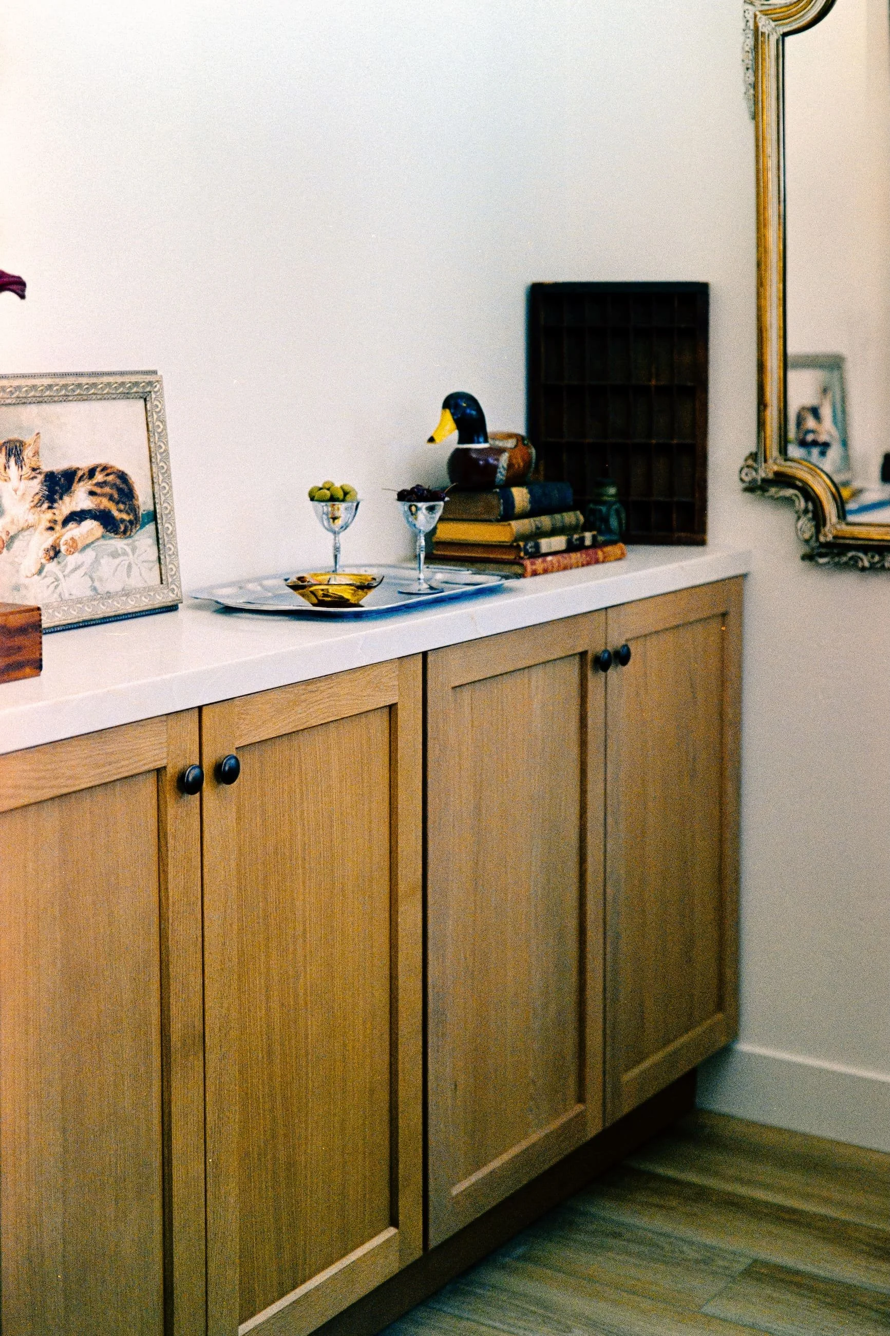 A wooden buffet with a white countertop, decorated with a framed picture of a cat, two glass bowls of fruit, and a ceramic duck figurine, with a small shelf and a mirror on the wall.