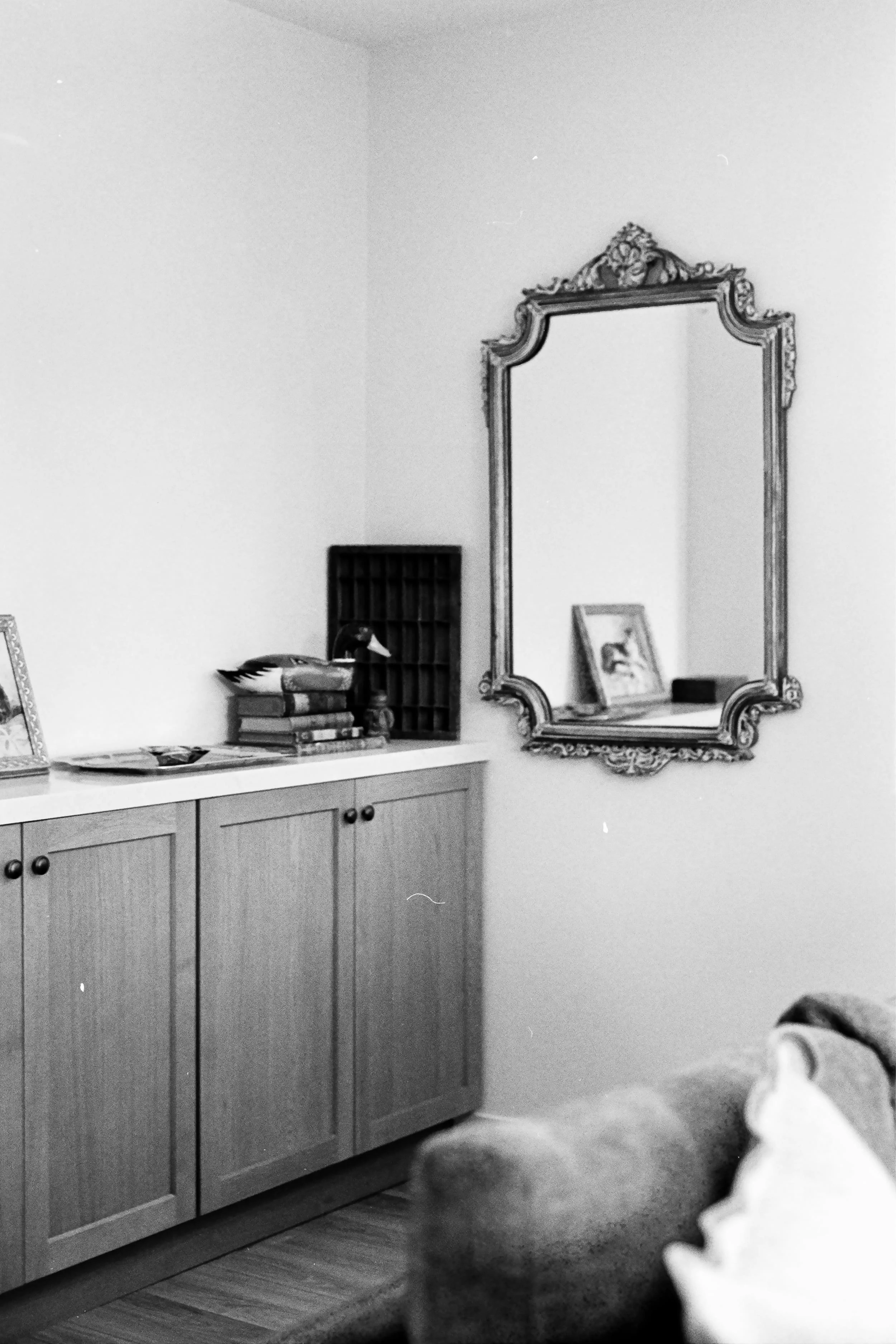 A black-and-white photo of a room corner featuring a wooden cabinet with closed doors, a stack of books, a small framed picture, and a decorative vintage mirror with intricate trim hanging on the wall.