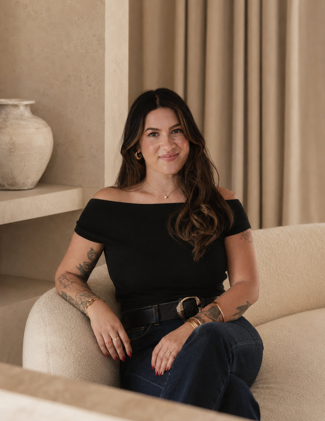 A woman with long dark hair and tattoos, wearing a black off-the-shoulder top, seated on a neutral-colored sofa in a modern, elegant room with beige walls and curtains.