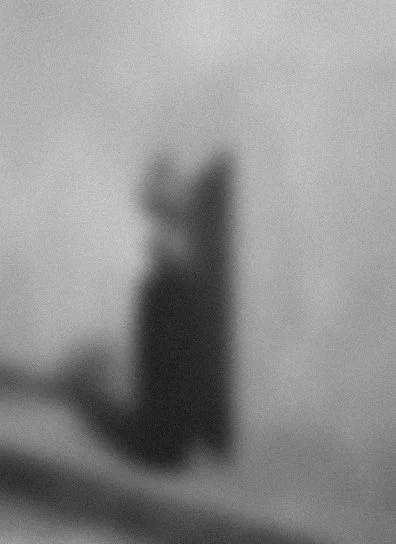 A blurry black cat sitting on a ledge against a plain wall.