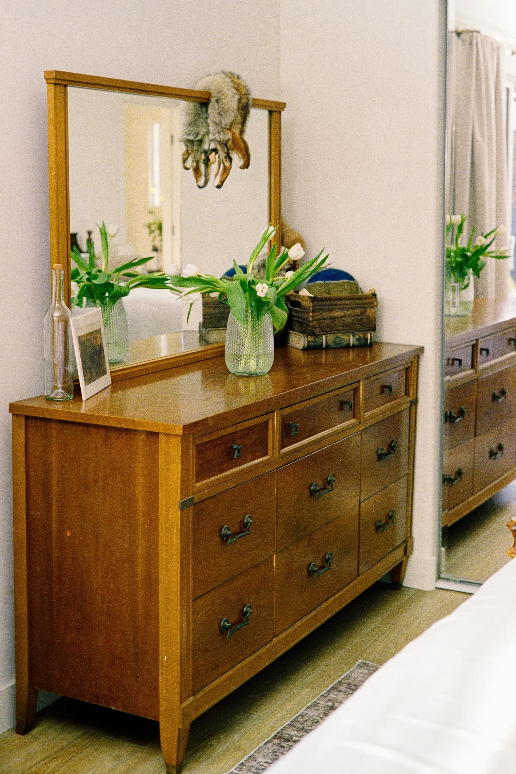 A wooden dresser with 12 drawers in a bedroom. On top of the dresser is a glass vase with white tulips, a small framed photo, and a decorative wooden box. A mirror is mounted on the wall above the dresser, reflecting part of the room including a stuffed animal hanging from the top of the mirror frame.