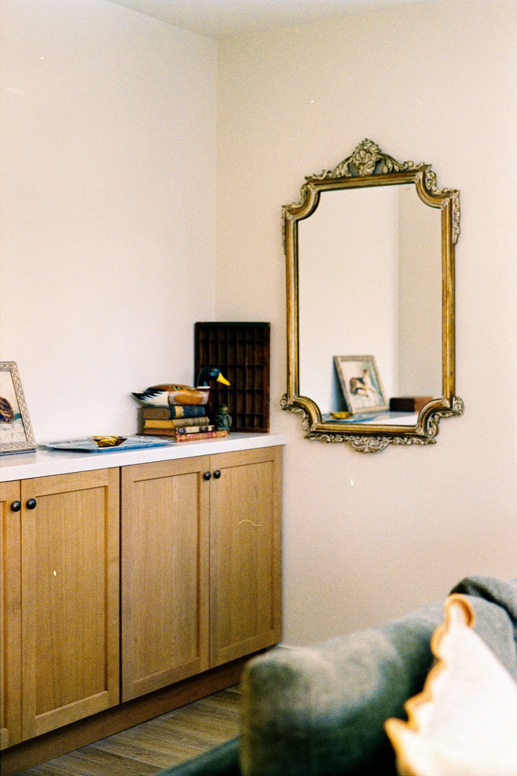 A beige cabinet with black knobs, topped with books, a decorative duck figure, and a framed picture, is in front of a white wall with a large ornate mirror hanging on it. There is a small dark-colored cabinet or shelf behind the decorative items.