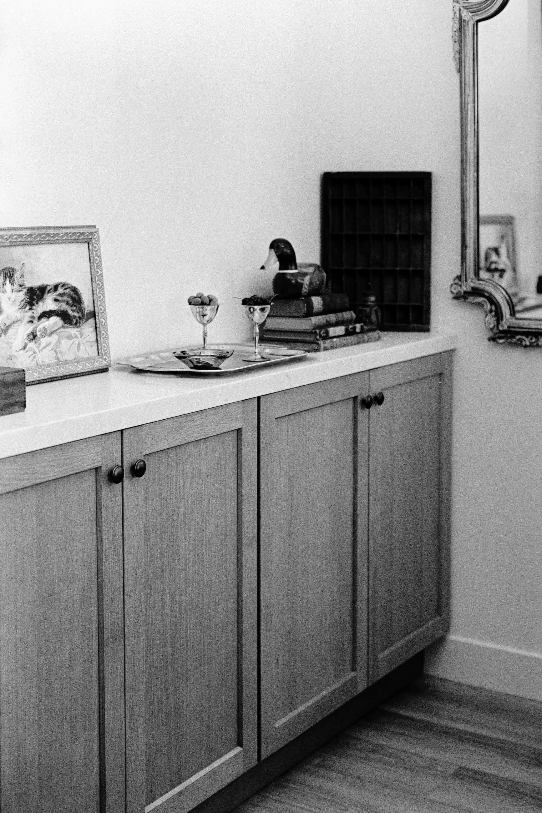 A wooden sideboard with decorative handles, topped with a framed picture of a cat, two glasses with fruit, a small dish, a set of books with a duck figurine on top, a small cubby shelf, a mirrored wall piece, and a hardwood floor.