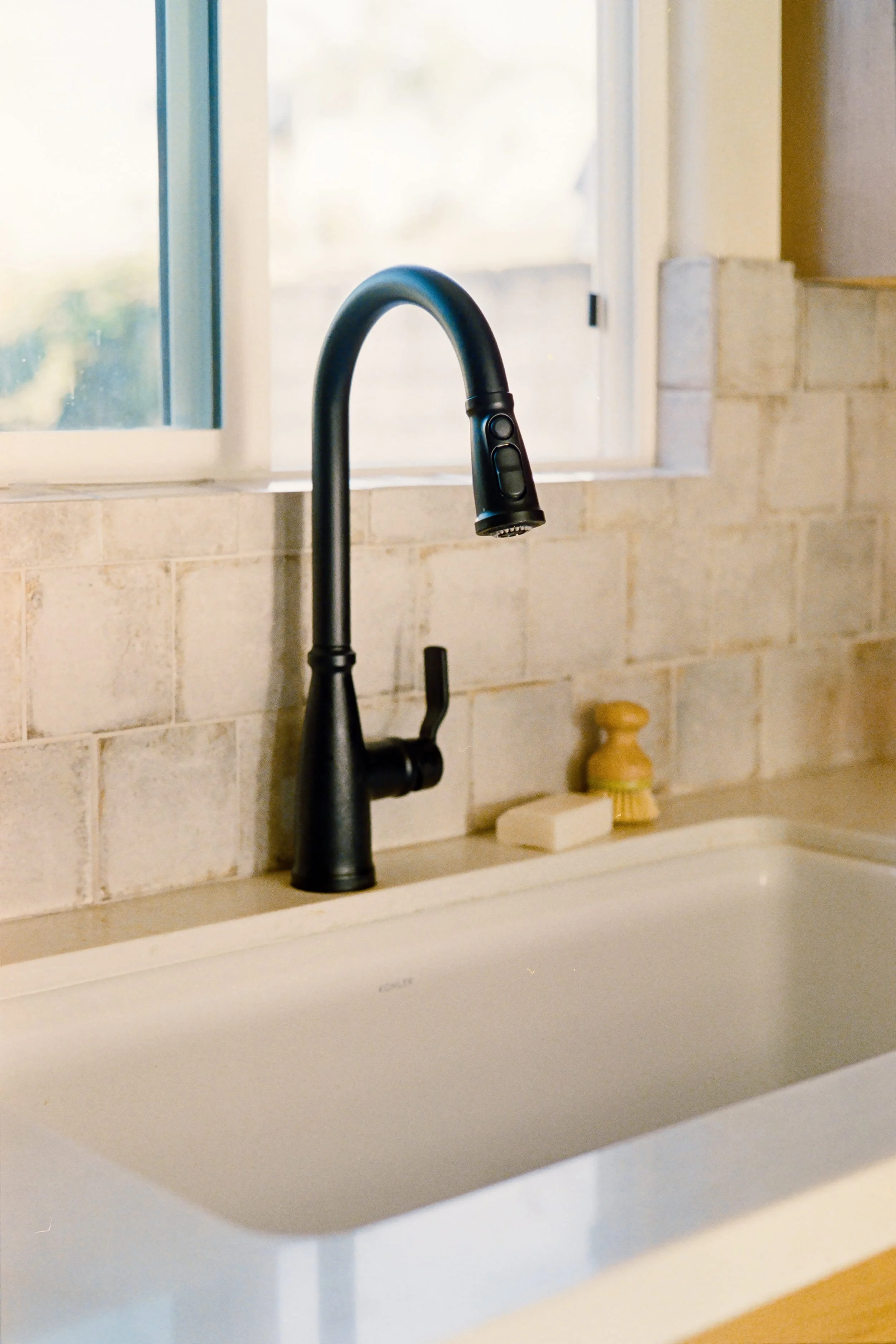 Close-up of a black kitchen faucet above a white sink, against a beige tiled backsplash and a window in the background.