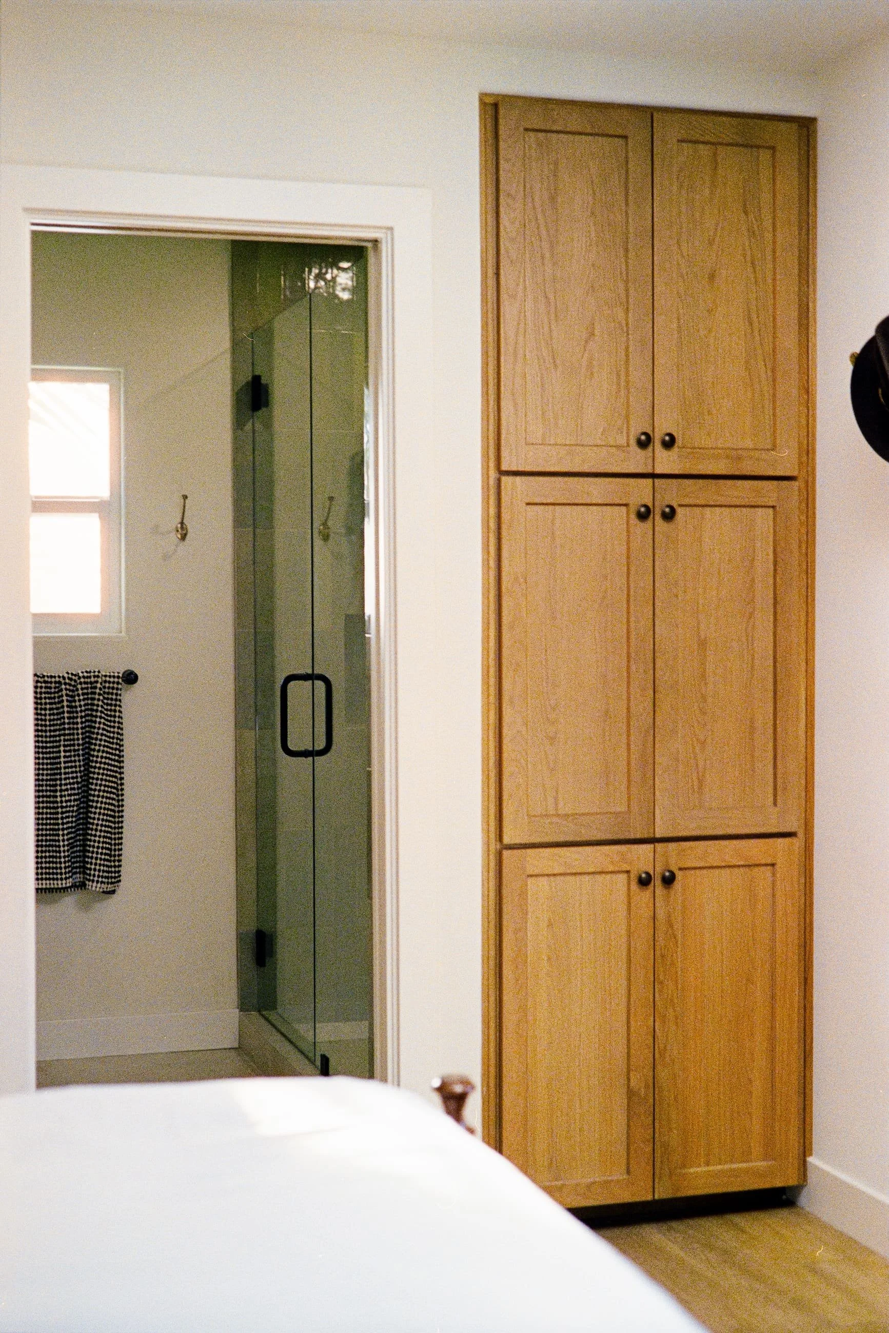 A bedroom with a white bed, a wooden cabinet with four doors, a glass shower door, a towel with a checkered pattern, a window, and hardwood flooring.