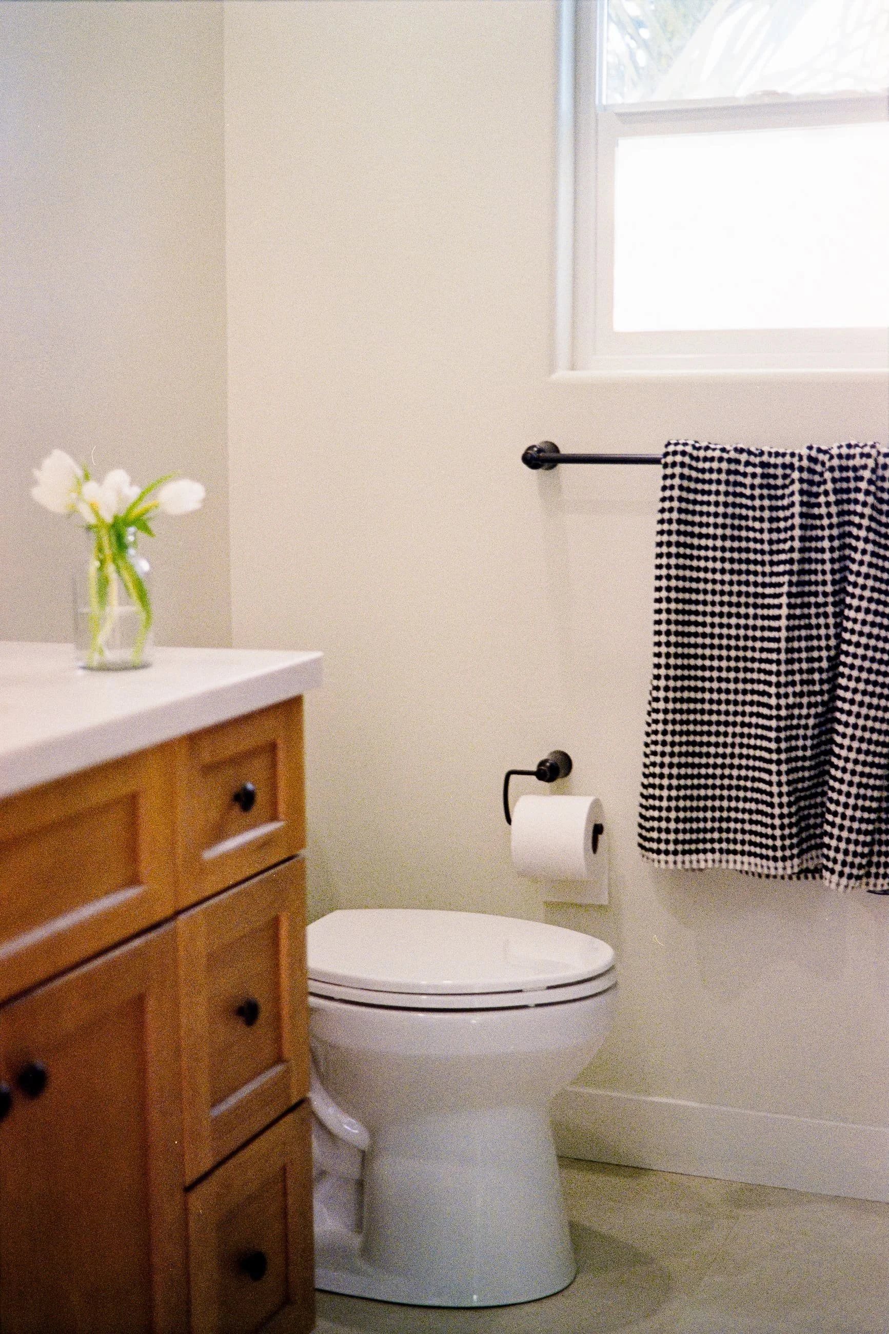 A bathroom with beige walls, a wooden vanity with black knobs, a small glass vase with white flowers on the counter, a window with a black curtain rod and a checkered towel hanging, and a white toilet with a closed lid and toilet paper on a roller.