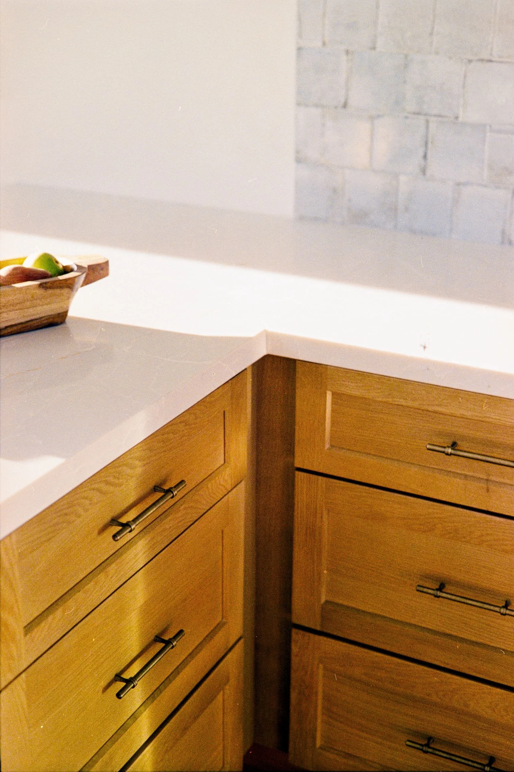 Kitchen corner with wooden cabinets and a white countertop. A fruit bowl with apples and bananas is on the countertop. Light-colored brick wall in the background.