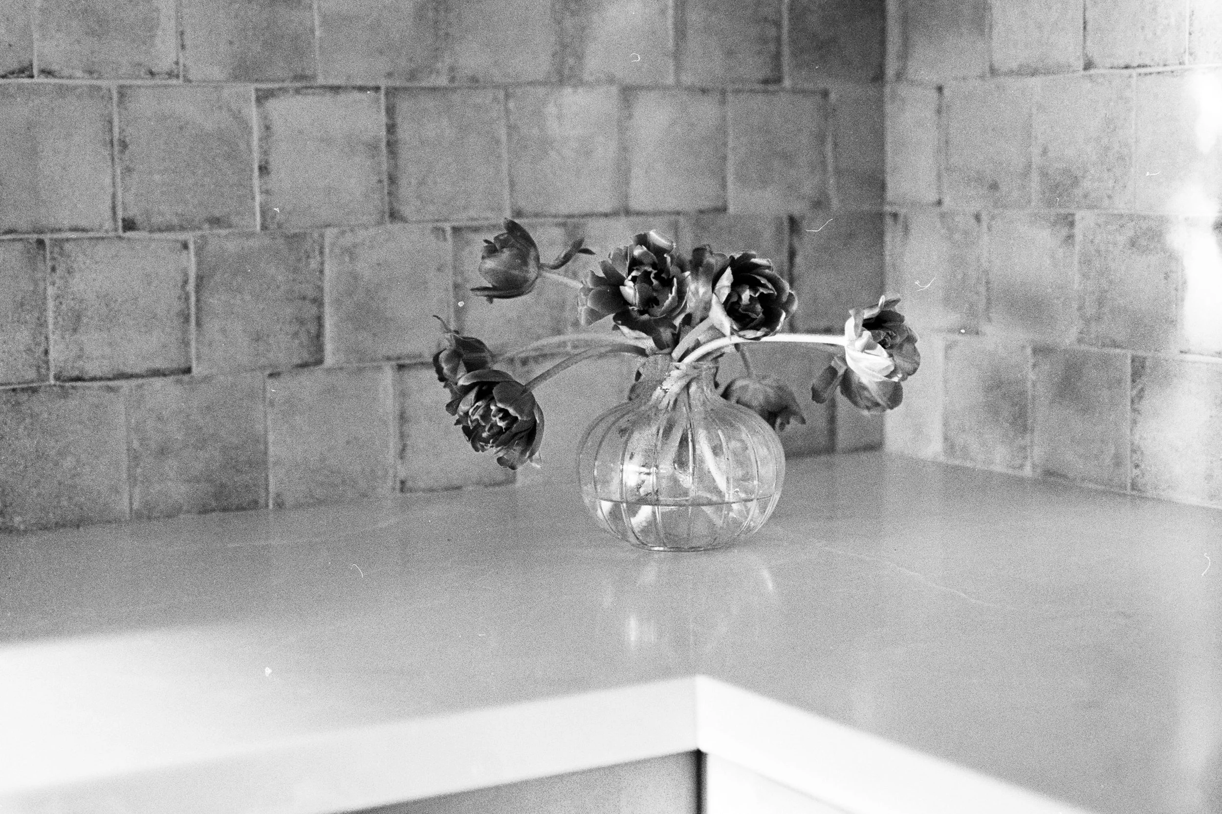 A black-and-white photo of a glass vase with wilted flowers on a table, against a brick wall background.
