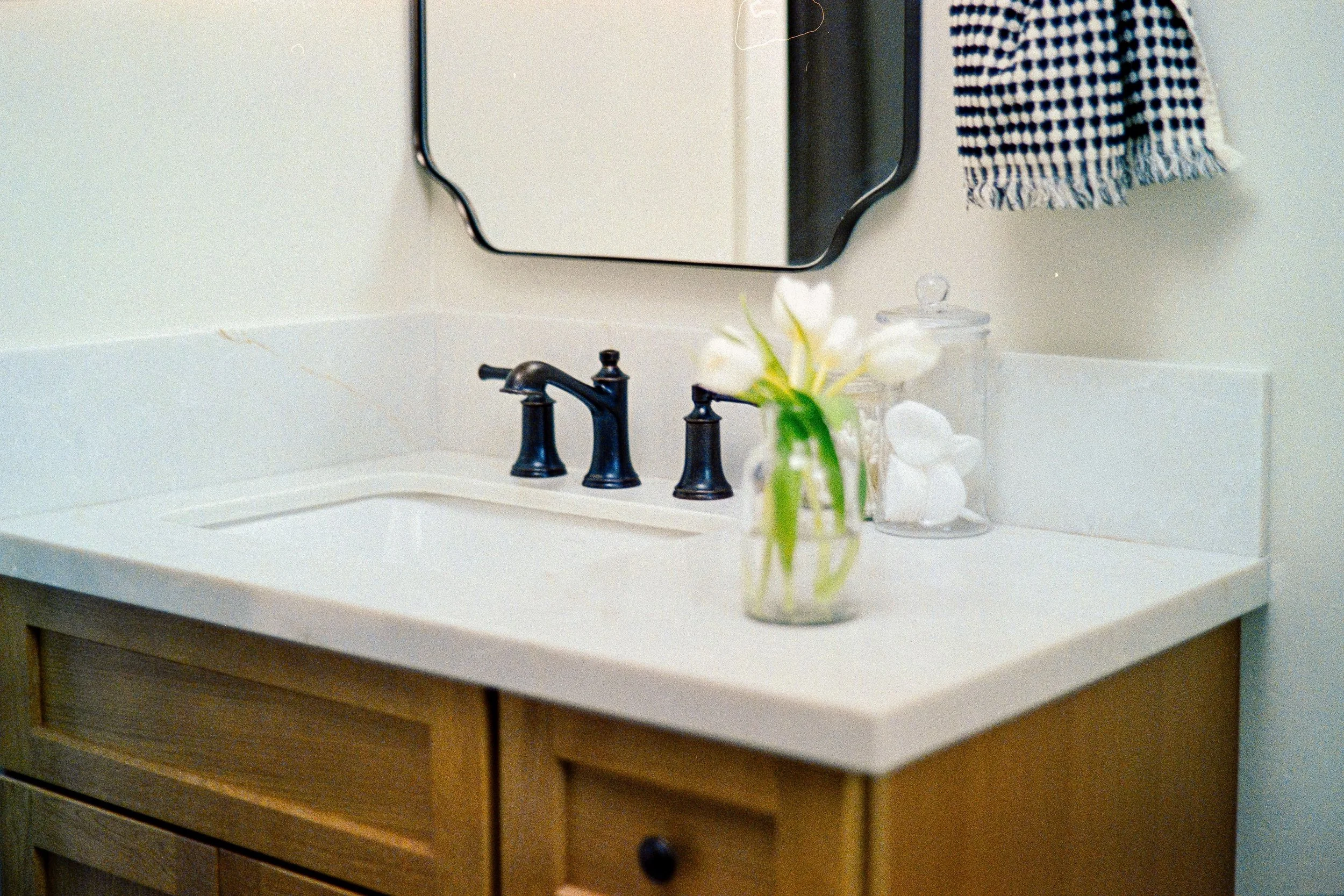 A bathroom vanity with a white countertop, a black faucet, and a mirror above. There is a glass jar with white flowers and a salt lamp next to it. A towel with a blue and white pattern is hanging on the wall.