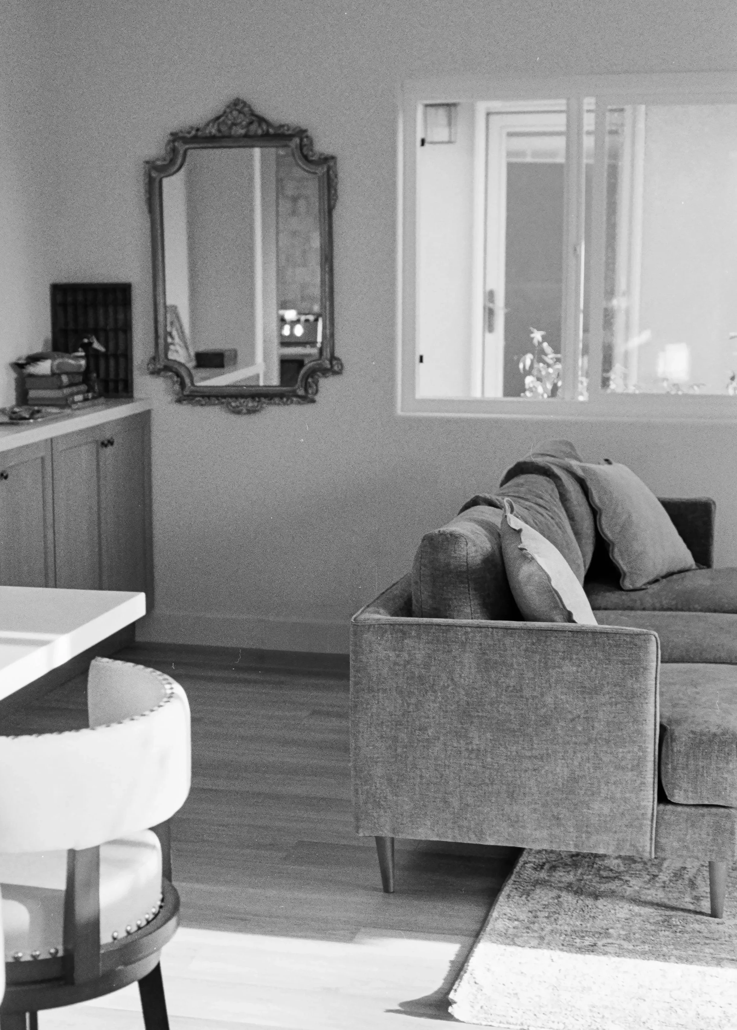 A living room with a large ornate mirror on the wall, a window letting in natural light, a sofa with cushions, a small white table, and a partial view of a white chair in the foreground.