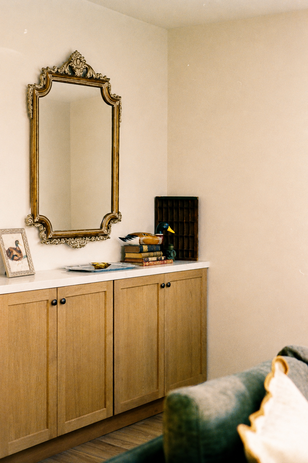 A wooden sideboard with a white top, decorated with a duck figurine, wooden books, and a small shelving unit, topped with an ornate gold mirror, against plain beige walls.