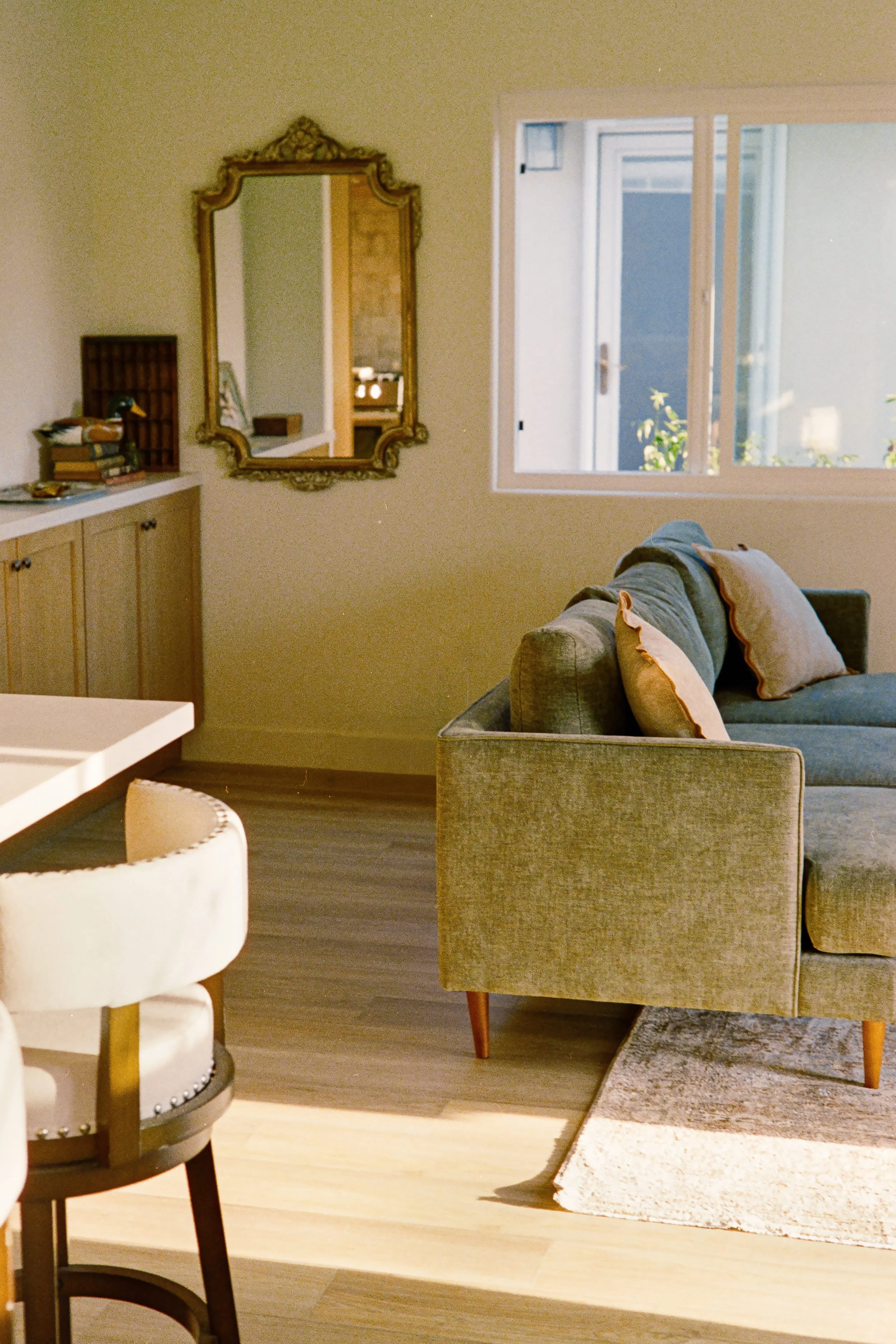 Living room with beige walls, a mirror, window, sofa with cushions, armchair, layered rug, and wooden flooring.