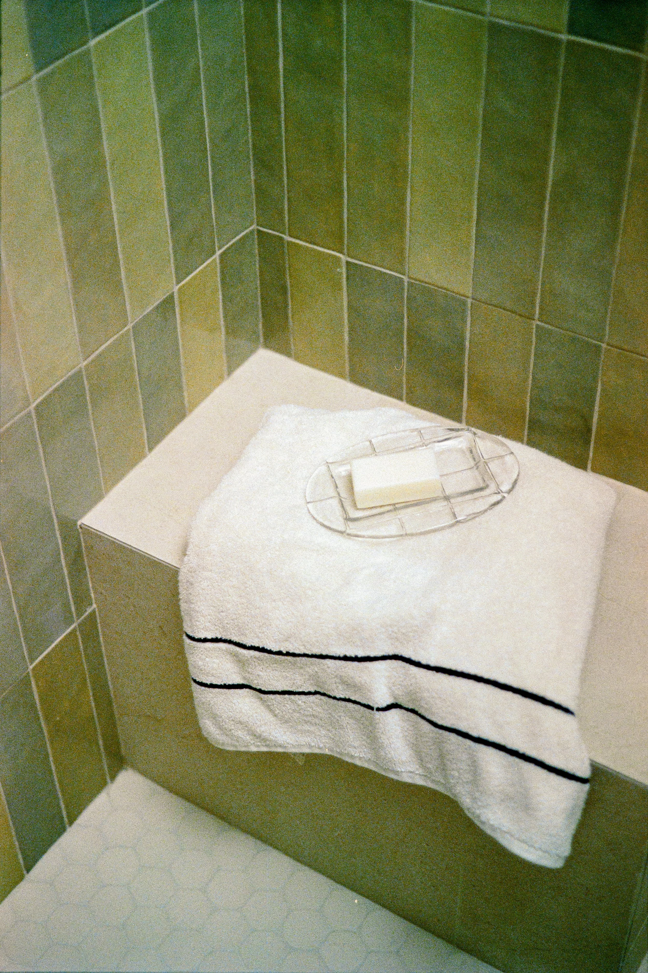 A bathroom corner with green tiled walls and a white hexagonal tiled floor. A small table with a white towel and a soap dish with a bar of soap is placed on the side of a dark-colored vanity or cabinet.