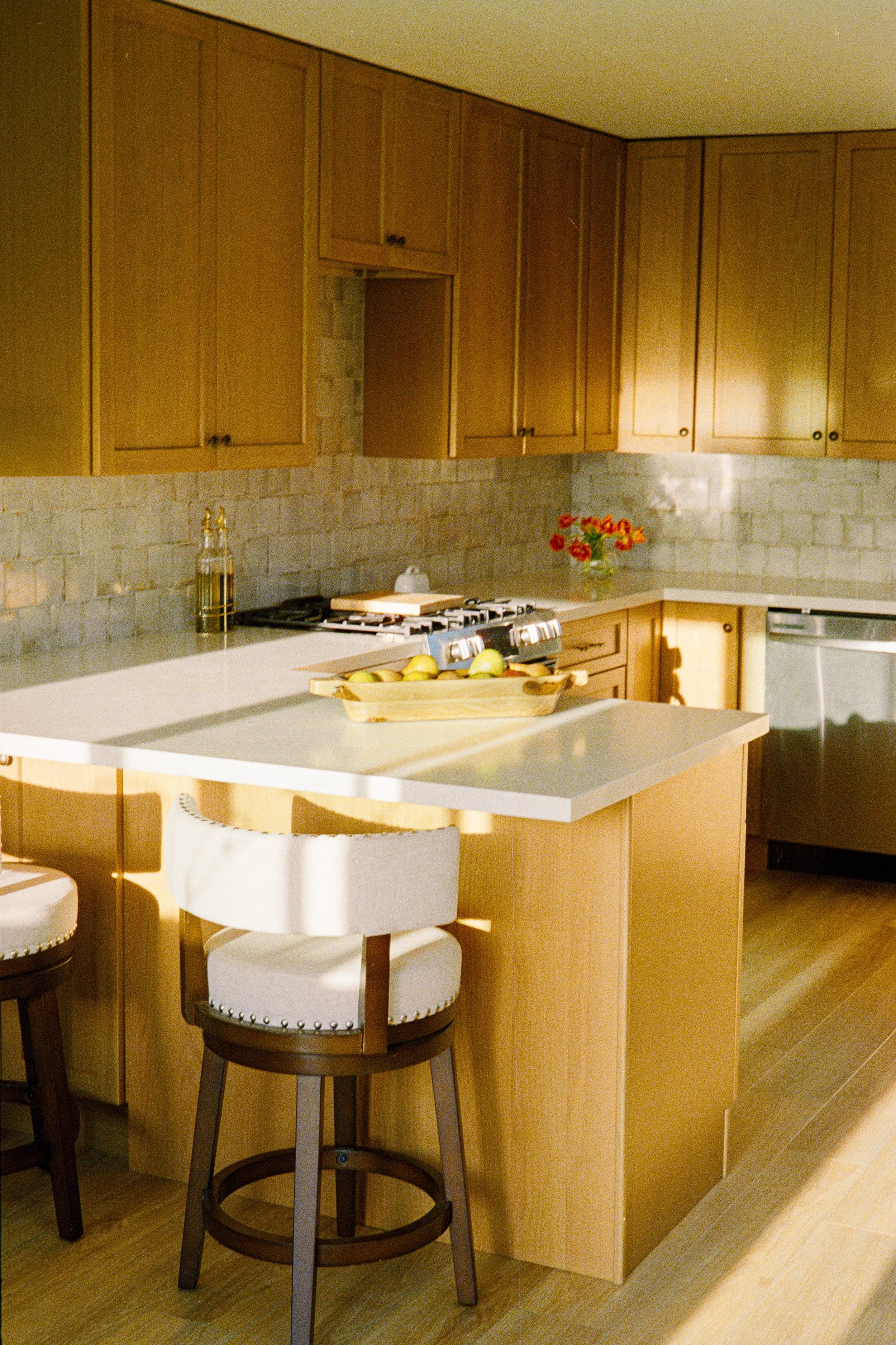 A kitchen with wooden cabinets, a gray brick backsplash, a bowl of green apples on the counter, a vase of red and orange flowers, a gas stove, a white kitchen chair, and sunlight casting shadows.