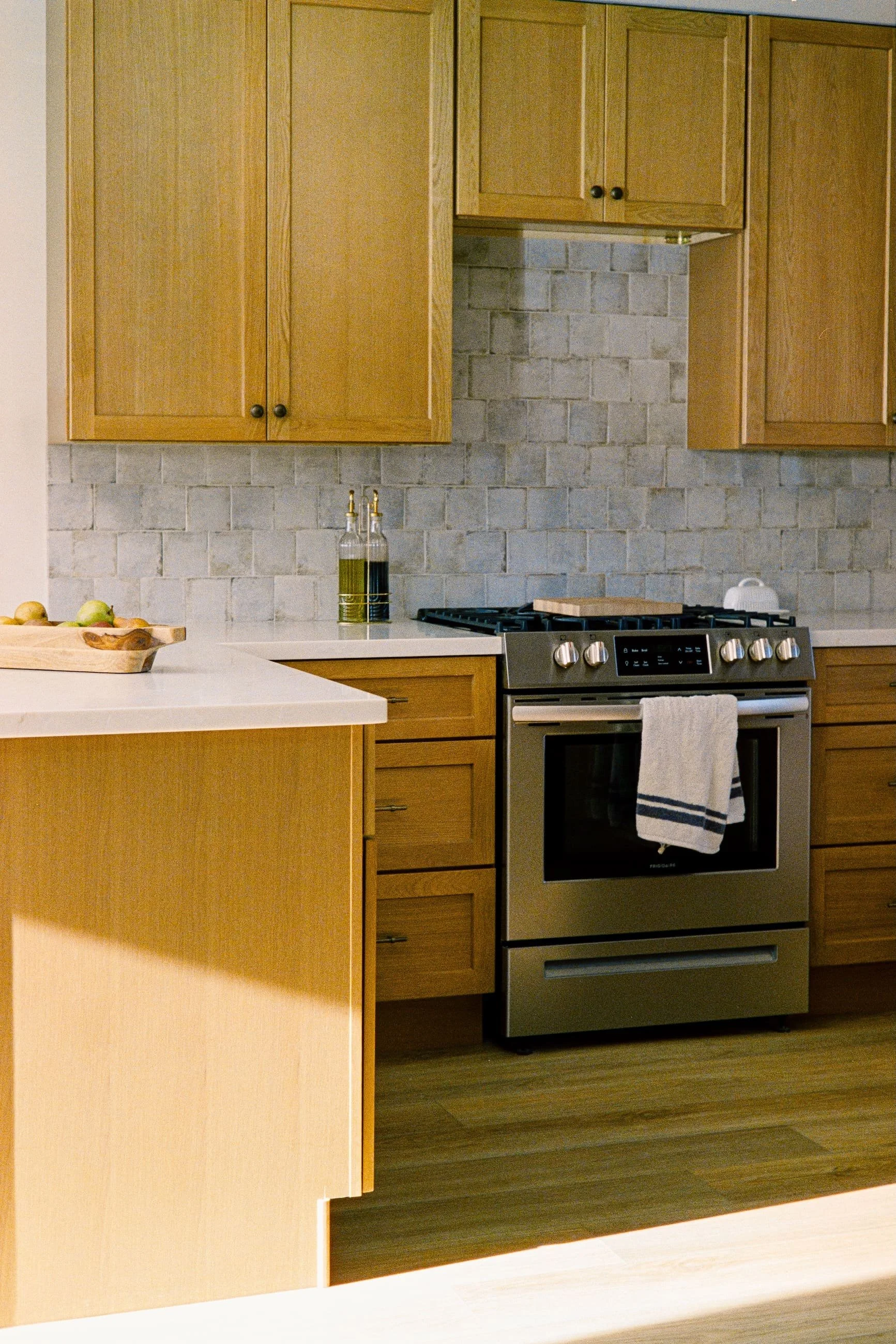 Modern kitchen with wooden cabinets, a gray brick backsplash, a stove, and a white countertop with a bowl of fruit.