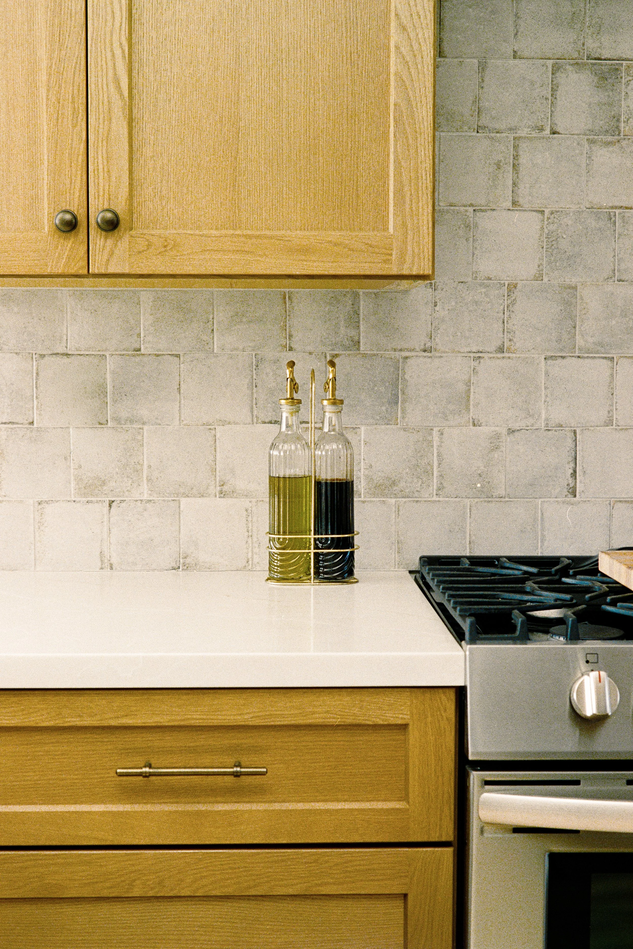 Kitchen with wooden cabinets, gray tiled backsplash, two glass bottles with oils, and a stove with black grates.
