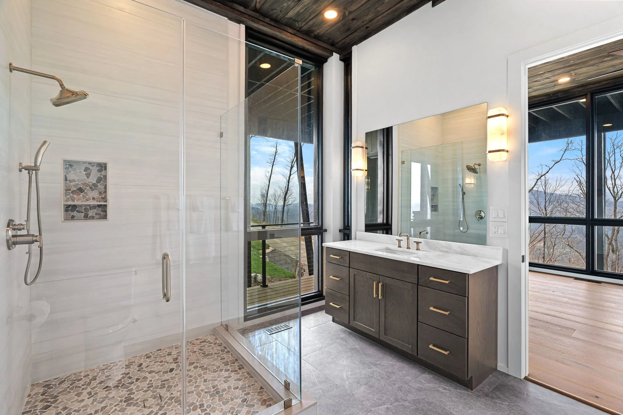 Modern bathroom with walk-in shower, large mirror, double sink, and wooden cabinets, with large windows showing an outdoor view of trees and sky.