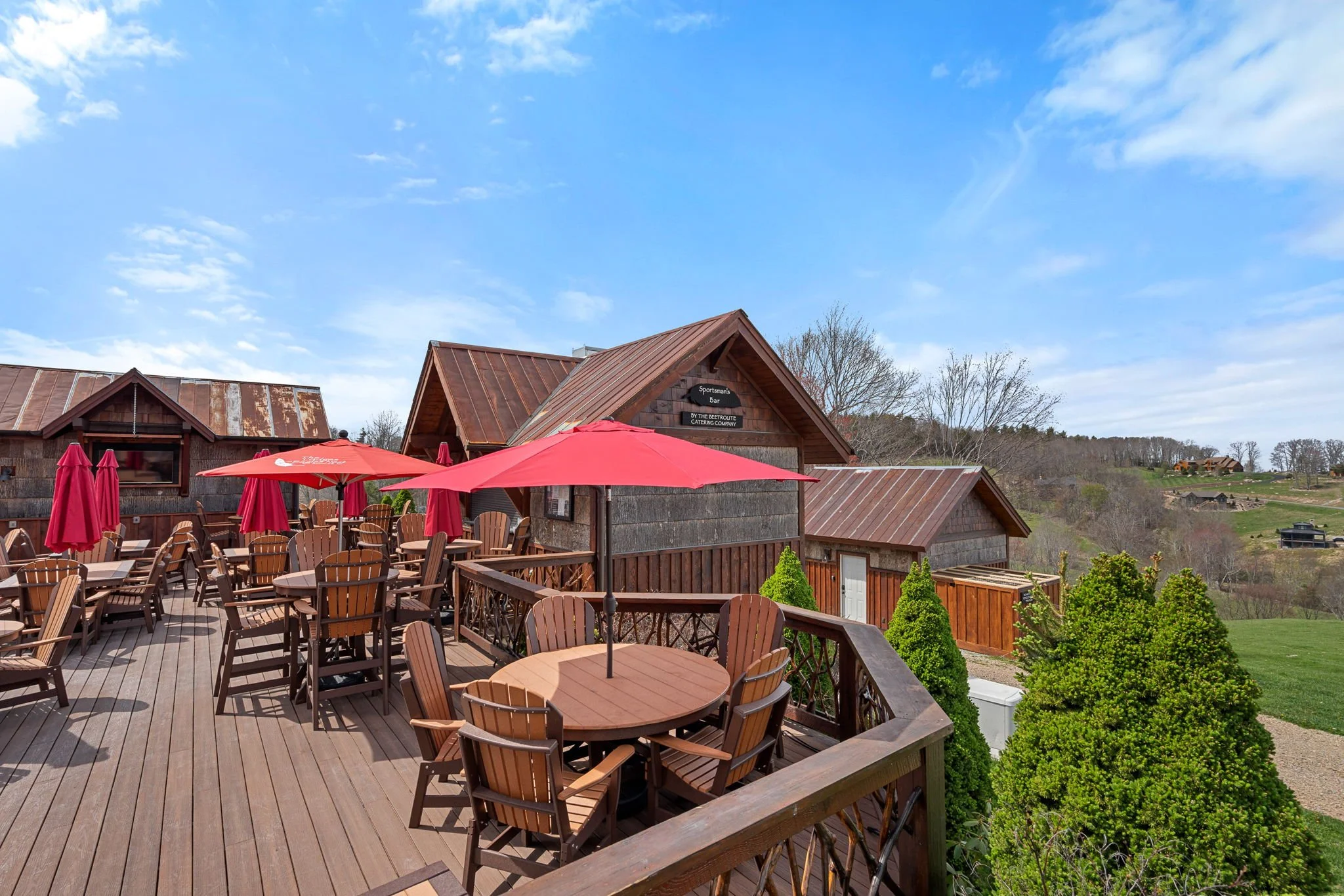 An outdoor restaurant or bar with wooden decking, red umbrellas, and wooden chairs on a sunny day with blue sky and scattered clouds. There are buildings with rustic, weathered metal roofs and greenery in the background.