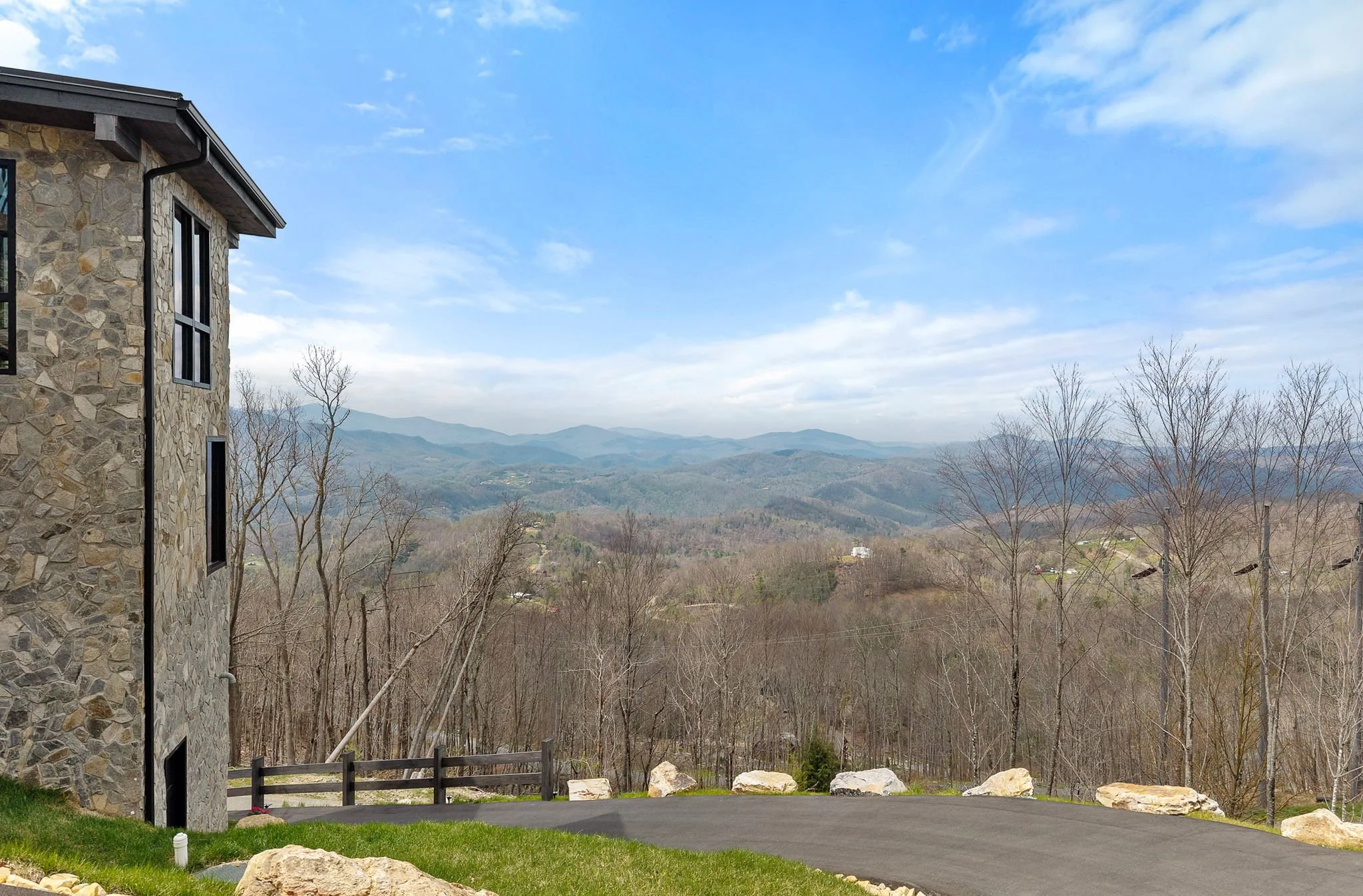 View of a mountain landscape with rolling hills and trees, taken from a house with stone exterior wall, large windows, and a paved driveway.