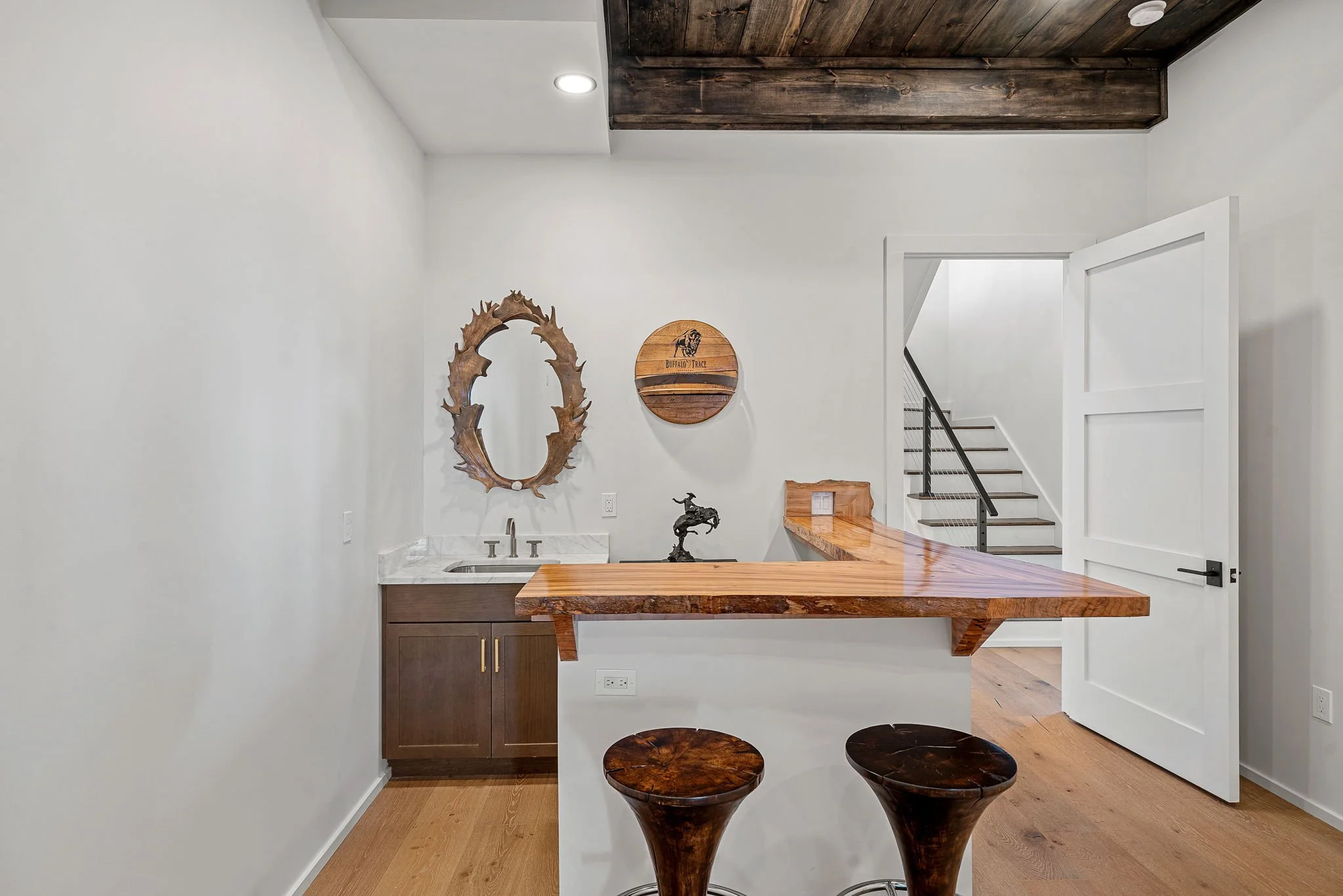 Modern kitchen area with white walls and a wooden breakfast bar with three matching wooden stools. Decorative items on the wall include a deer antler mirror and a circular wooden art piece with a lion logo and the words "Buffalo Trace." A staircase w