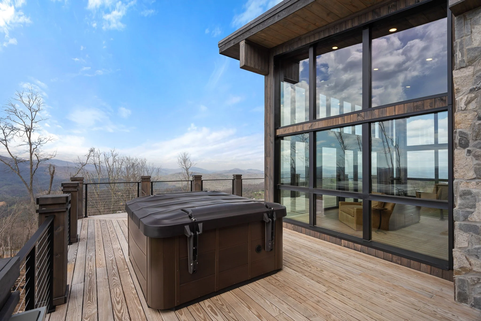 Modern balcony with wooden flooring, hot tub, glass windows reflecting cloudy sky, and mountain landscape in the background.