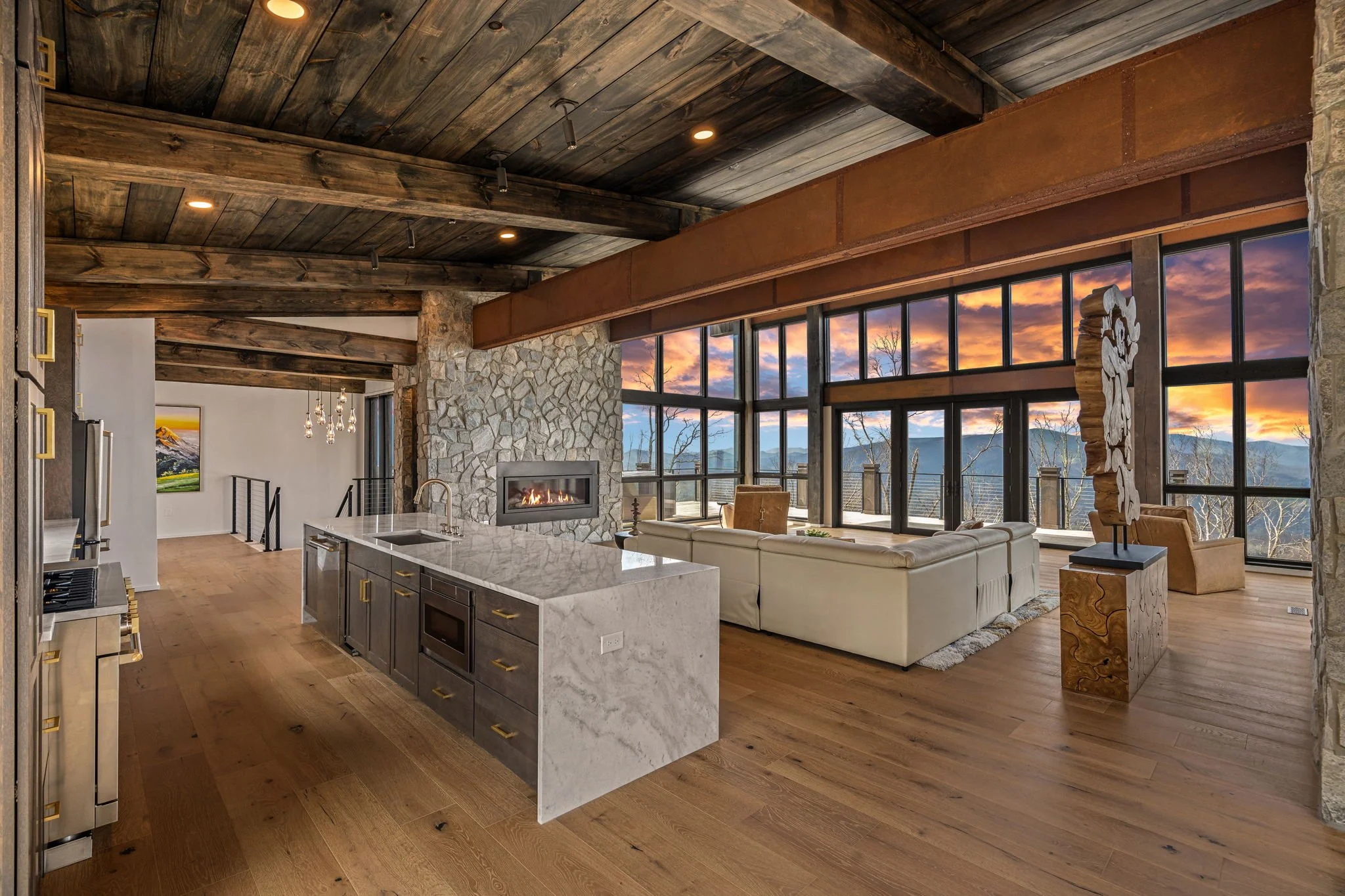 Modern living room with large windows showing a mountain view at sunset, stone fireplace, wooden ceiling beams, and a kitchen island.