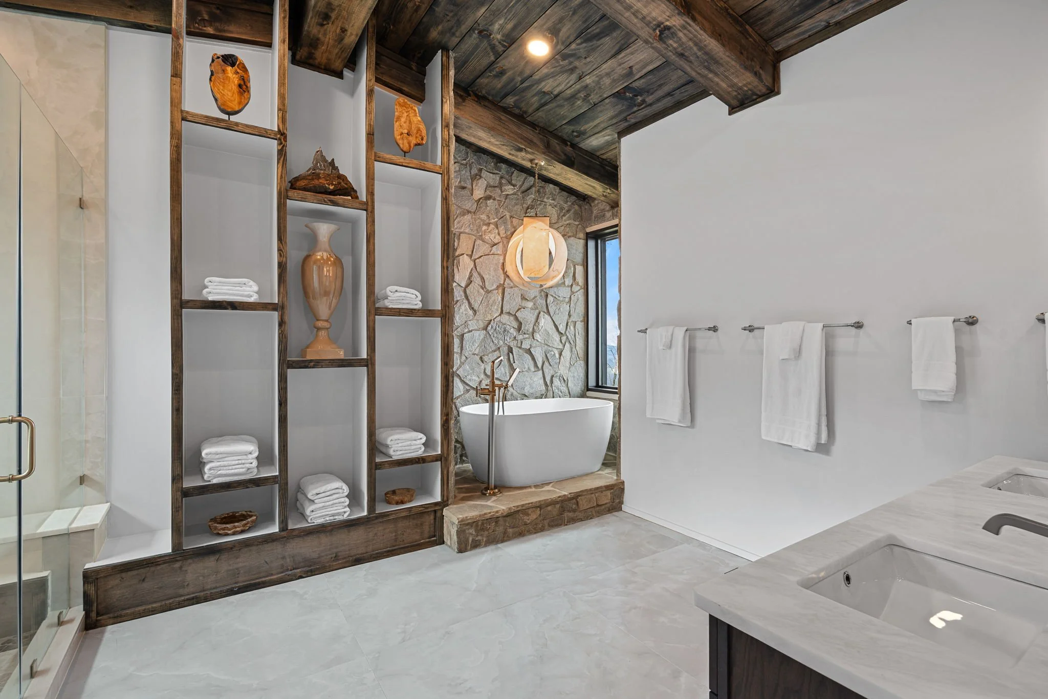 Modern bathroom with a white freestanding bathtub near a stone wall, a large window, wooden ceiling, and a white towel rack with towels. A wooden shelving unit holds towels and decorative items.