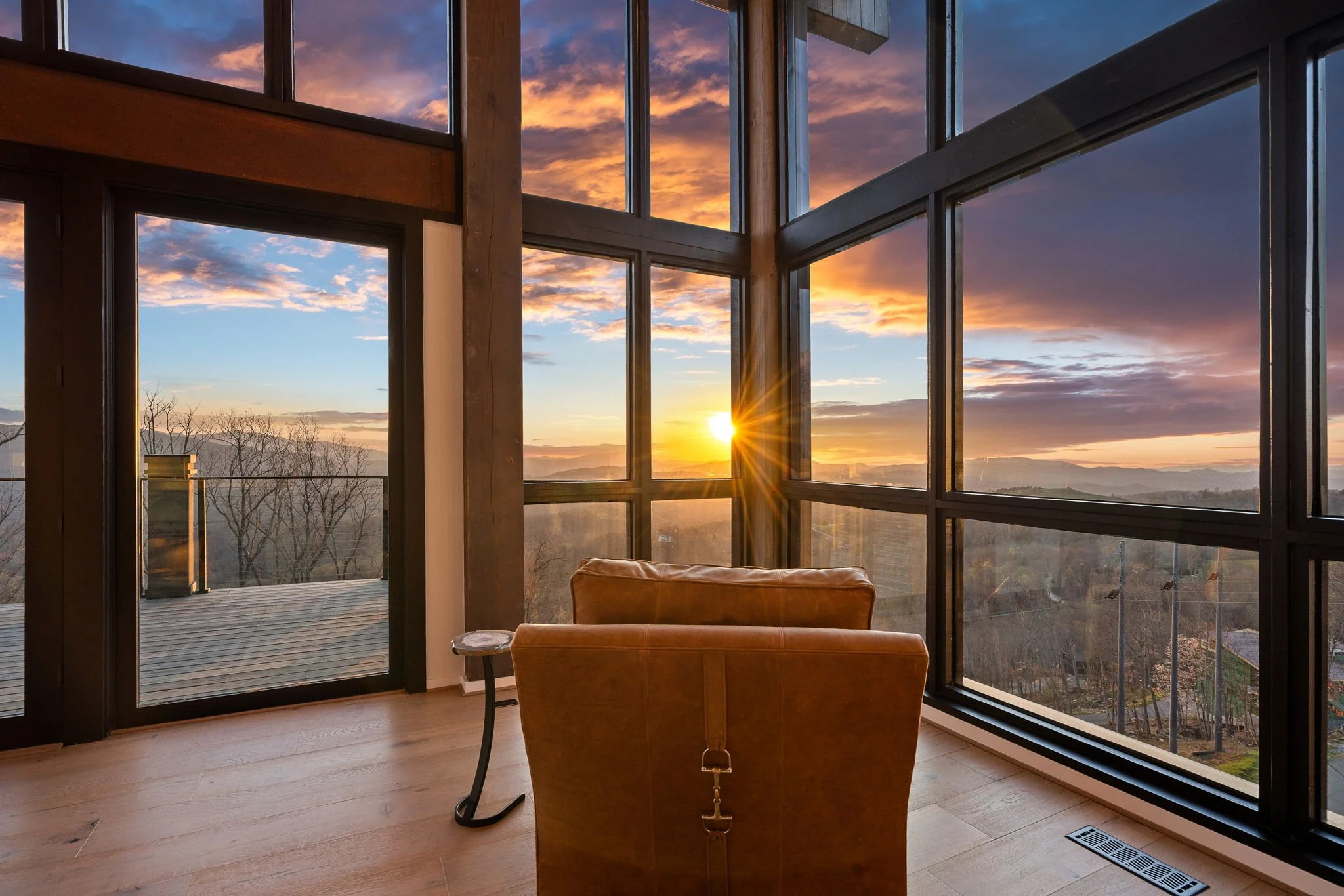 A living room with floor-to-ceiling windows showing a sunset over a mountain landscape. There is a brown leather chair and a small side table in the room, with a wooden deck visible outside.