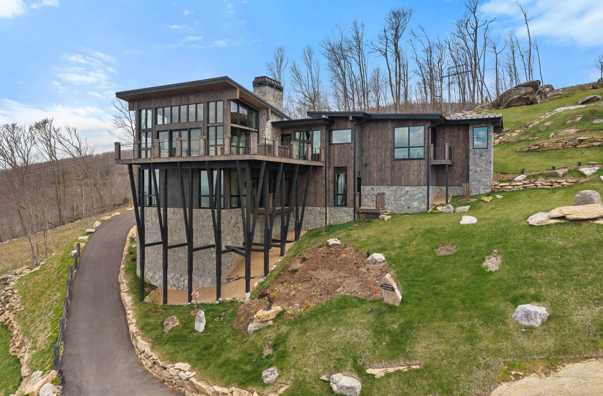 Modern house built into a hillside with a stone foundation, wooden siding, large glass windows, and a wooden deck, surrounded by grass, rocks, and leafless trees under a partly cloudy sky.