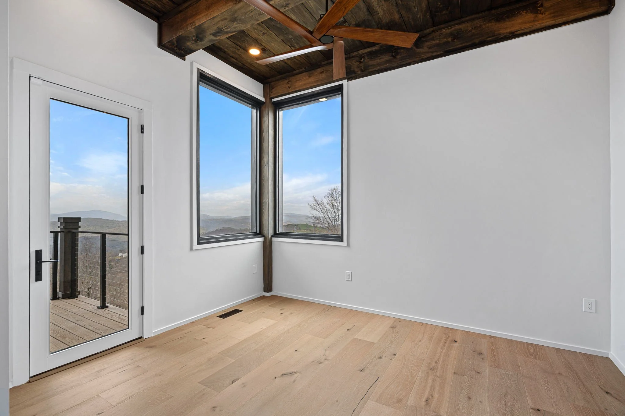 An interior room with white walls, large windows showing a landscape view, wooden ceiling with ceiling fan, and a door leading to an outdoor deck.