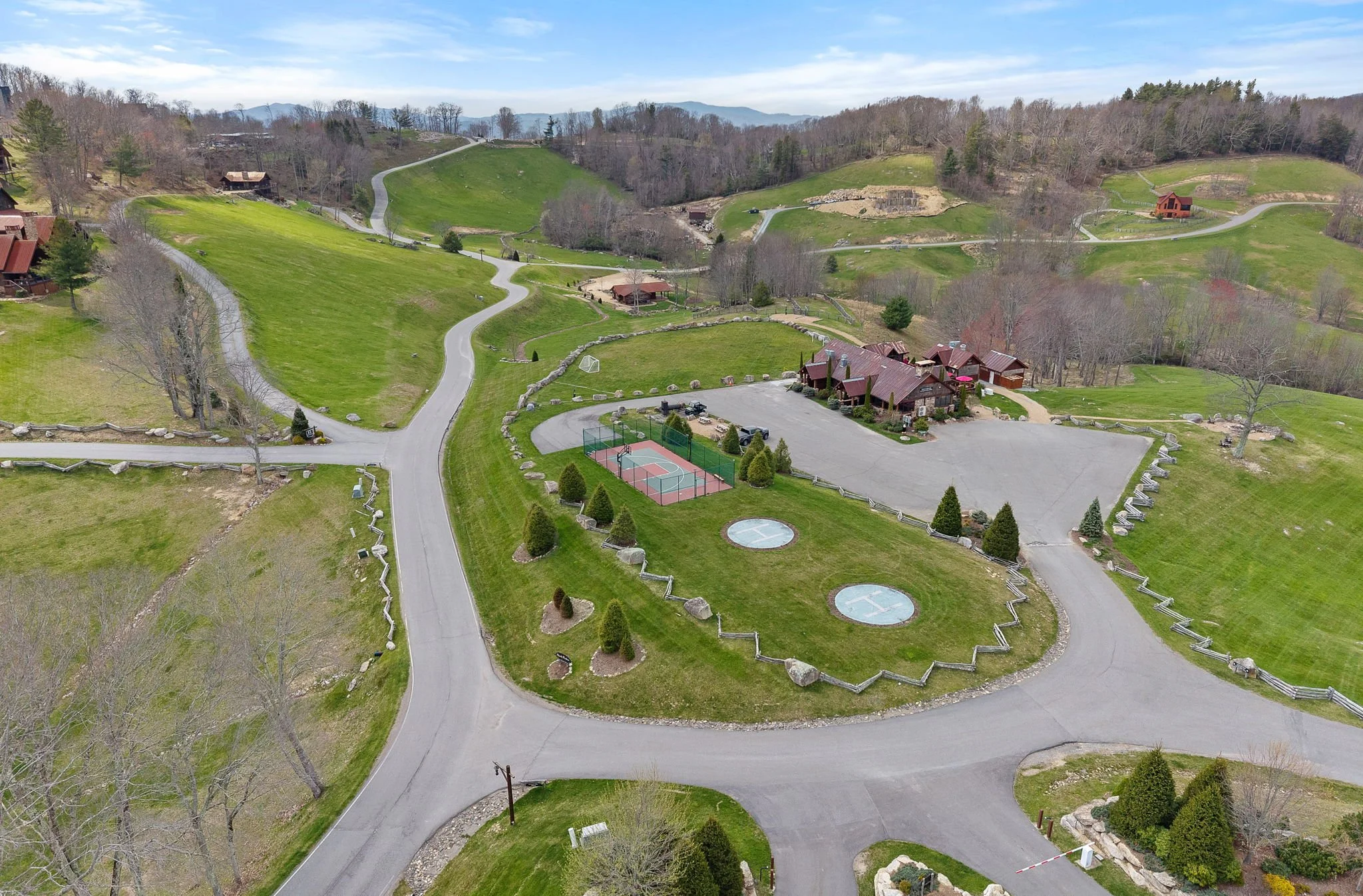 Aerial view of a green park area with winding paths, small buildings, tennis courts, and surrounding trees and hills under a partly cloudy sky.