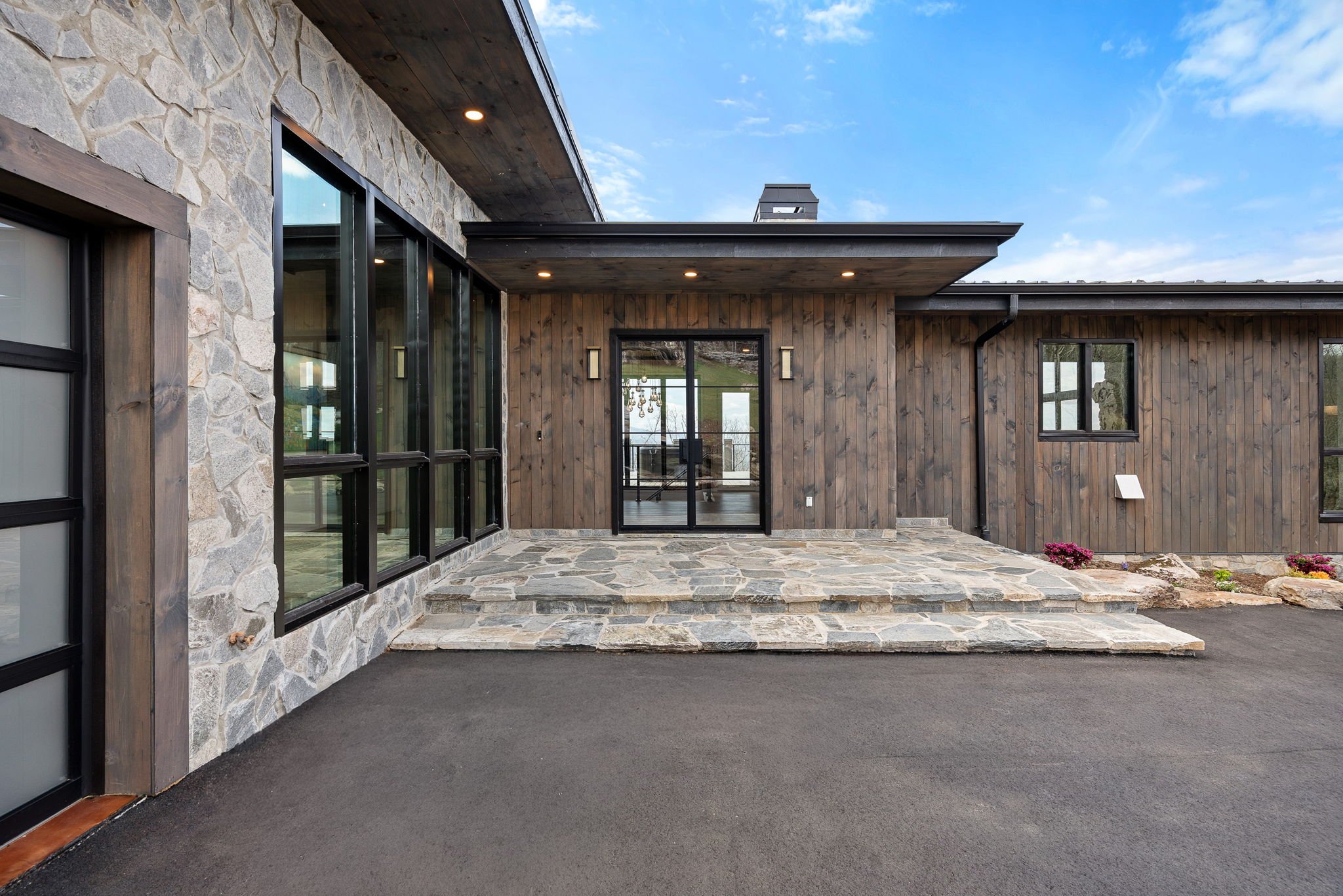 Modern house exterior featuring stone and wood siding, large windows, a glass door, stone steps, and a paved patio under a partly cloudy sky.