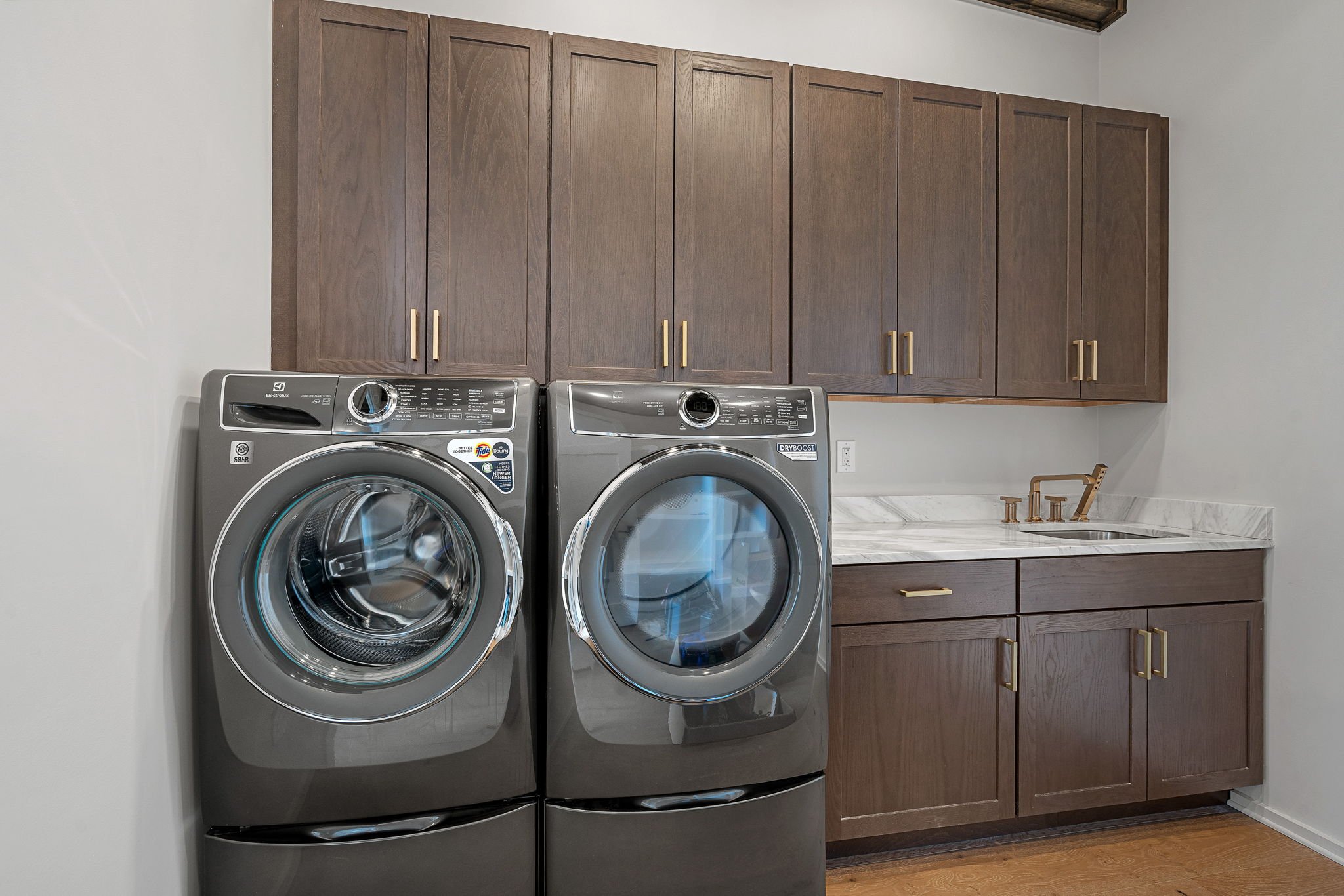 A laundry room with a front-loading washer and dryer in stainless steel finish, wooden cabinets above, a marble countertop, and a small gold-colored sink faucet on the right.