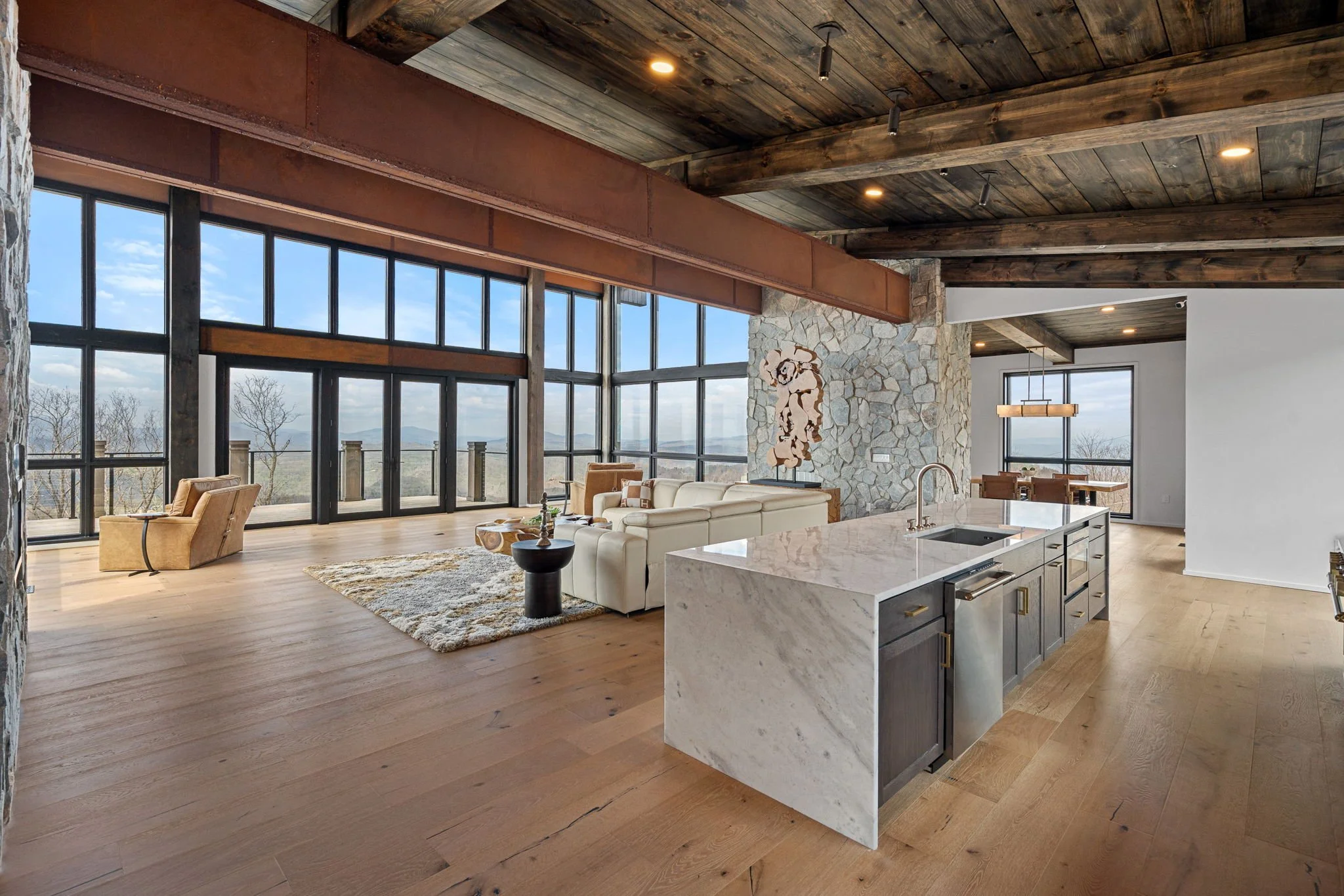 Open-concept living room and kitchen with large windows showing an outdoor landscape, wooden ceiling beams, stone wall, white sectional sofa, chairs, carpet, and a marble kitchen island.
