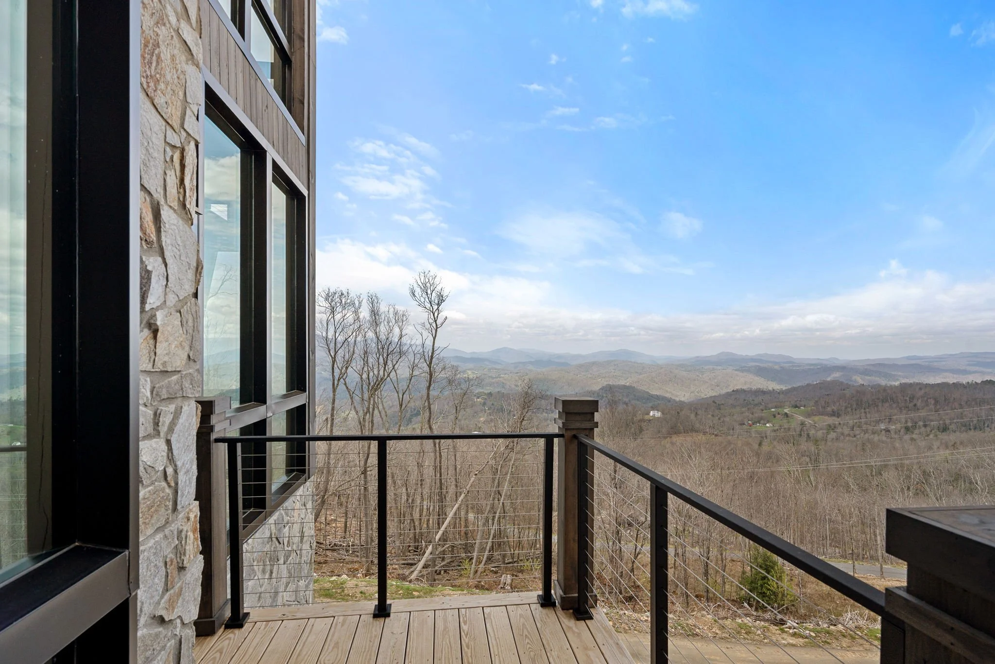 A balcony with a metal railing overlooking a landscape of leafless trees, hills, and mountains under a partly cloudy blue sky, adjacent to a building with stone and wood siding and large windows.