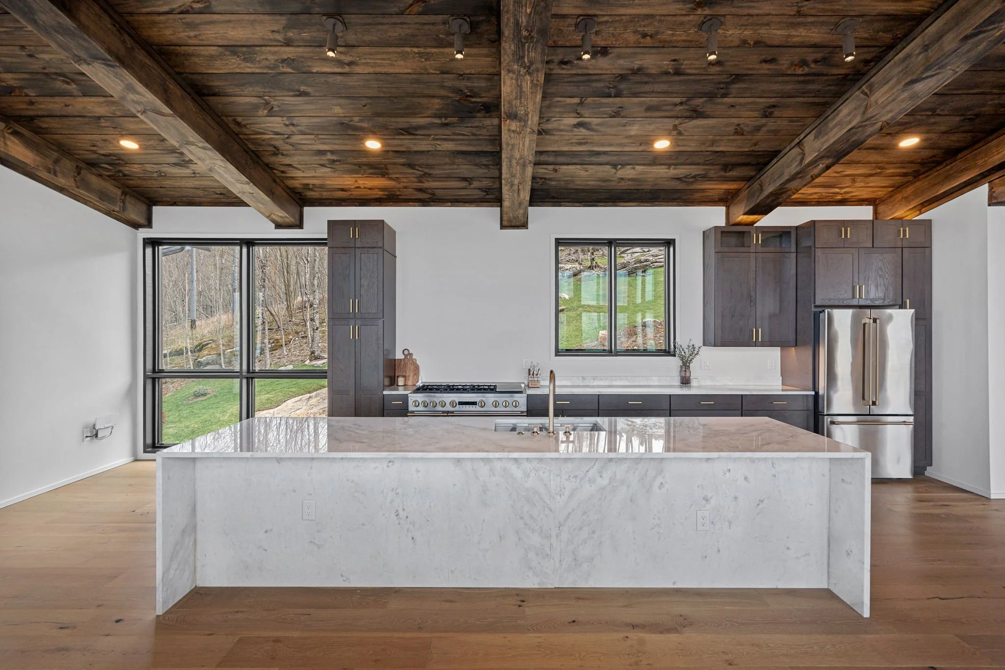 Modern kitchen with white marble island, dark gray cabinets, large windows showing outdoor greenery, and a wooden ceiling.