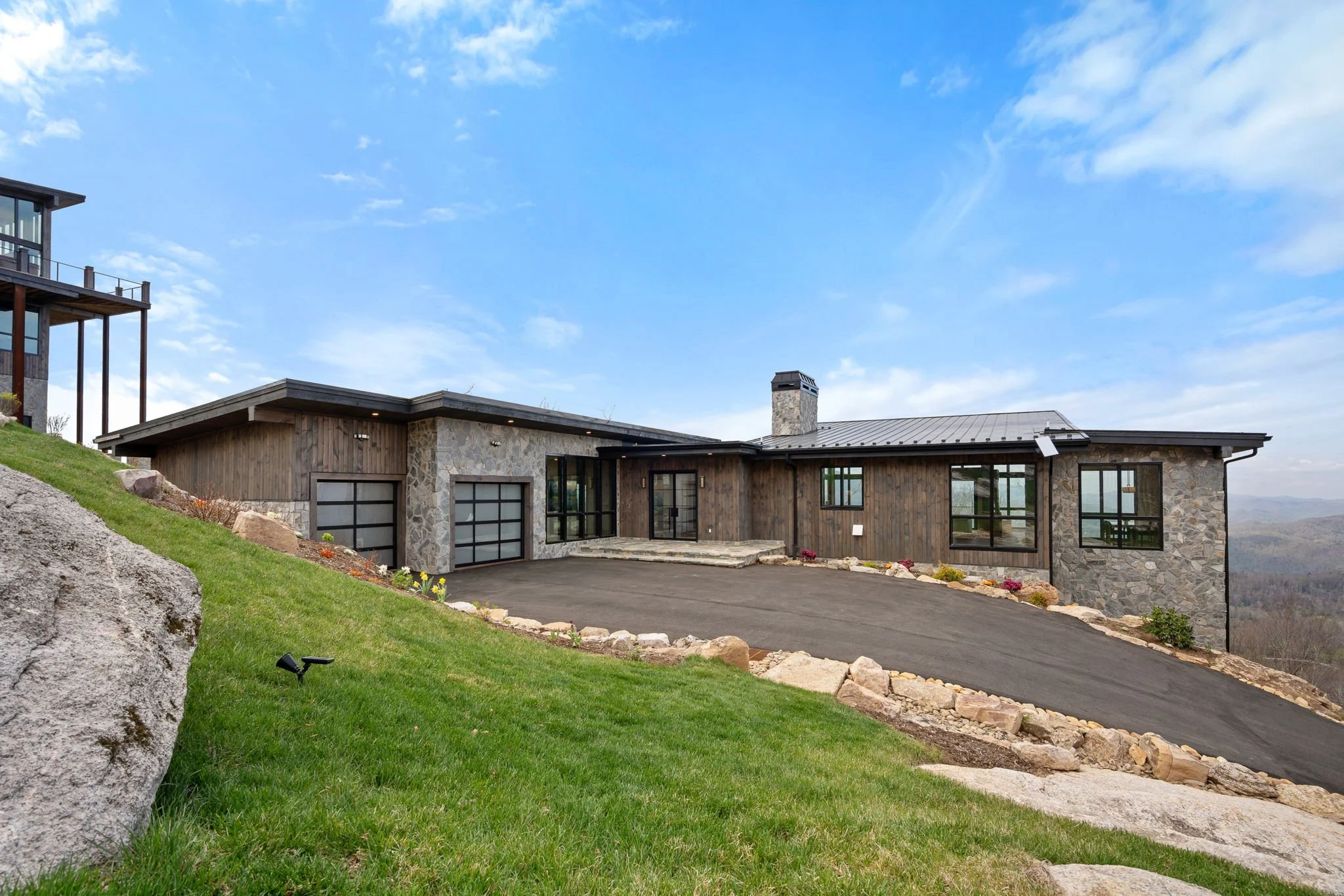 Modern house with stone and wood exterior, large windows, garage, black roof, chimney, and a driveway on a landscaped lawn with rocks and flowers under a blue sky with clouds.