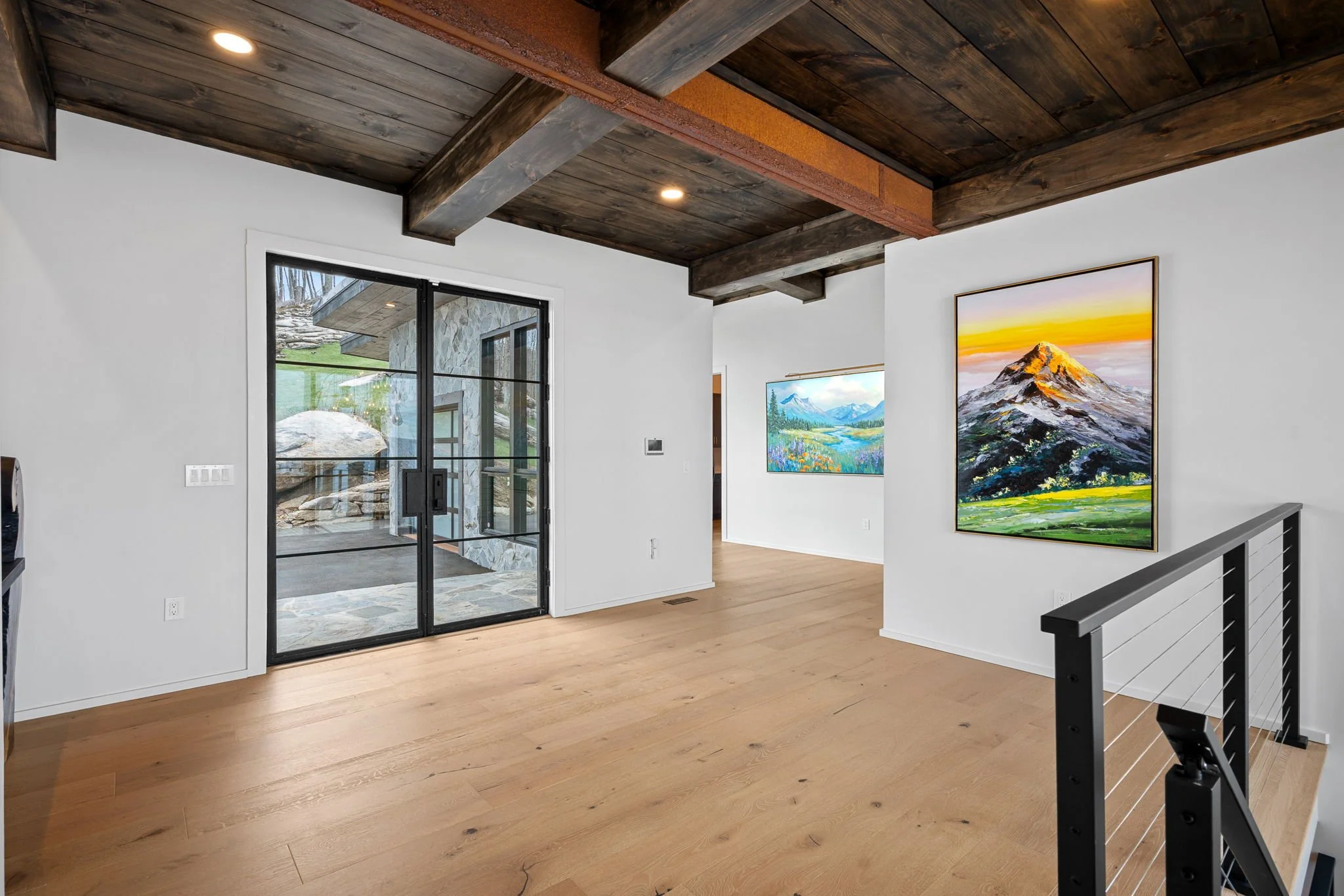 Interior of a modern home with a wooden ceiling, hardwood floors, and contemporary artwork on the walls, including mountain landscapes, near a glass door leading outside.