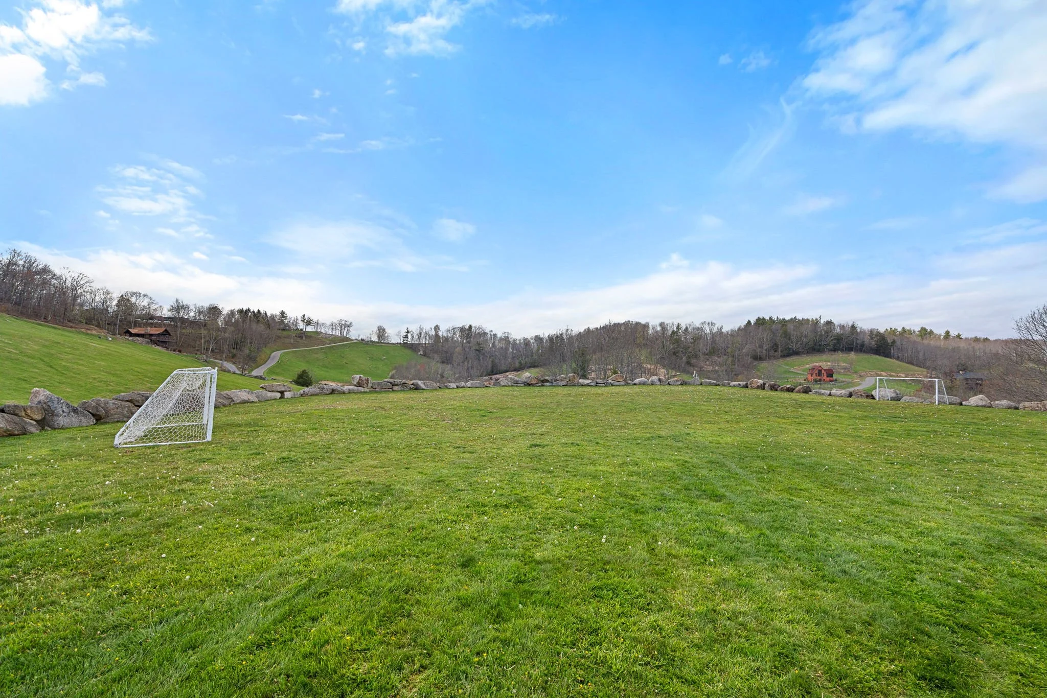 A grassy sports field with two soccer goals, surrounded by a stone border, and a scenic hillside with trees and houses under a partly cloudy blue sky.
