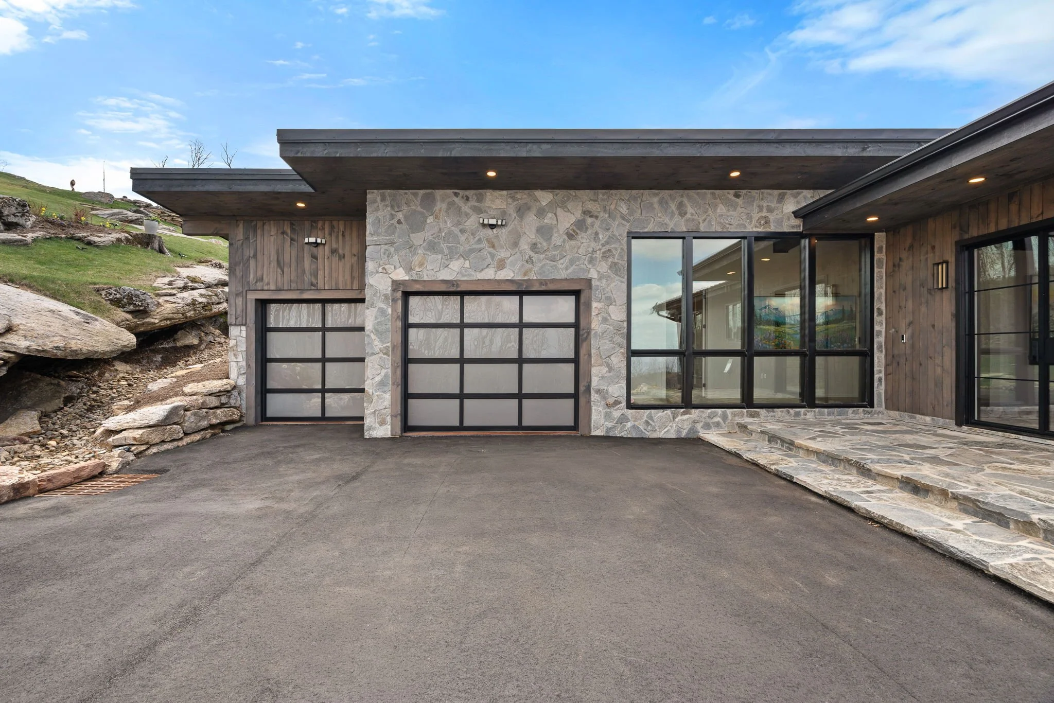 Contemporary house with stone and wood exterior, large garage door, glass windows, and a driveway, set against a hillside under a blue sky.