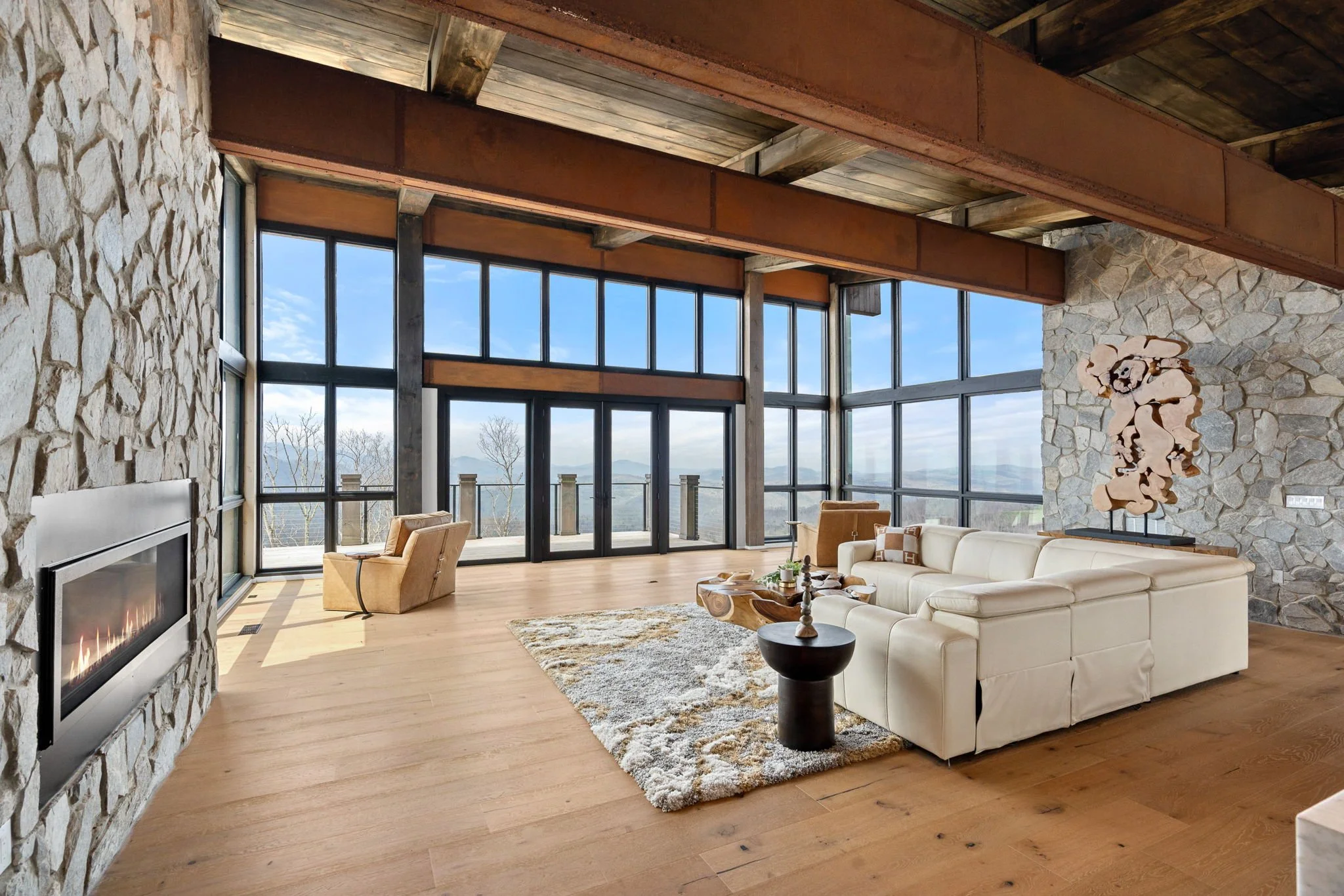Living room with large windows, stone walls, wooden beams, and beige furniture including a sectional sofa and armchairs.