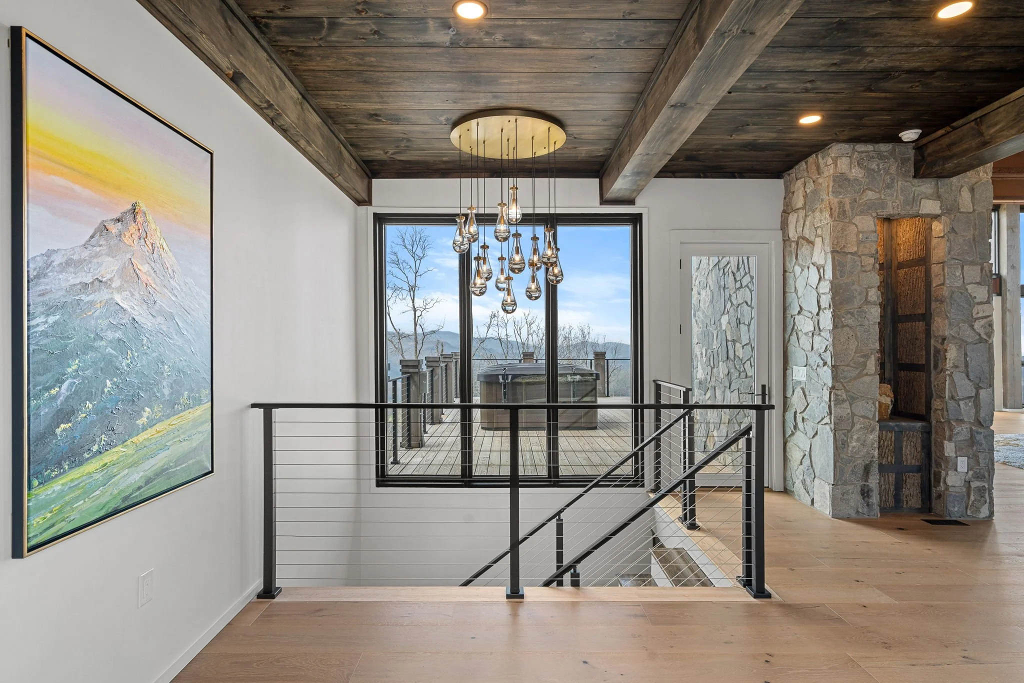 Interior view of a modern home with a staircase, large window, and a stone accent wall. The ceiling is wooden with recessed lighting, and a pendant light fixture with multiple bulbs hangs above the space. There is a landscape painting on the wall to 
